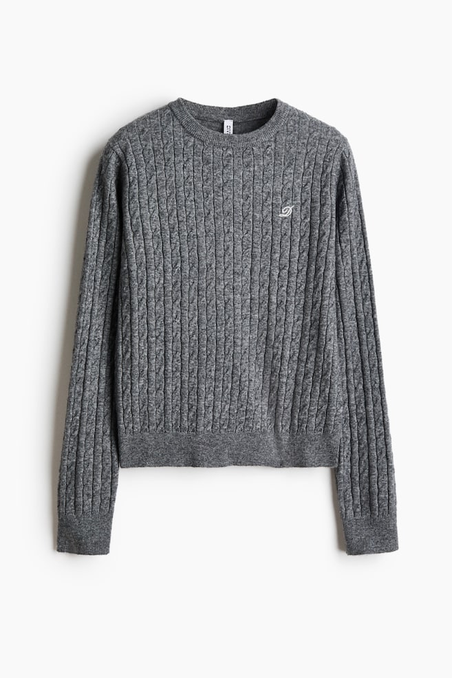 Un pull gris en maille torsadée est présenté de face, mettant en valeur un col rond côtelé, des poignets côtelés et un ourlet côtelé. Le pull présente un motif vertical en maille torsadée sur lâensemble du corps et des manches, avec un petit monogramme brodé blanc détaillé sur la poitrine gauche. - 1