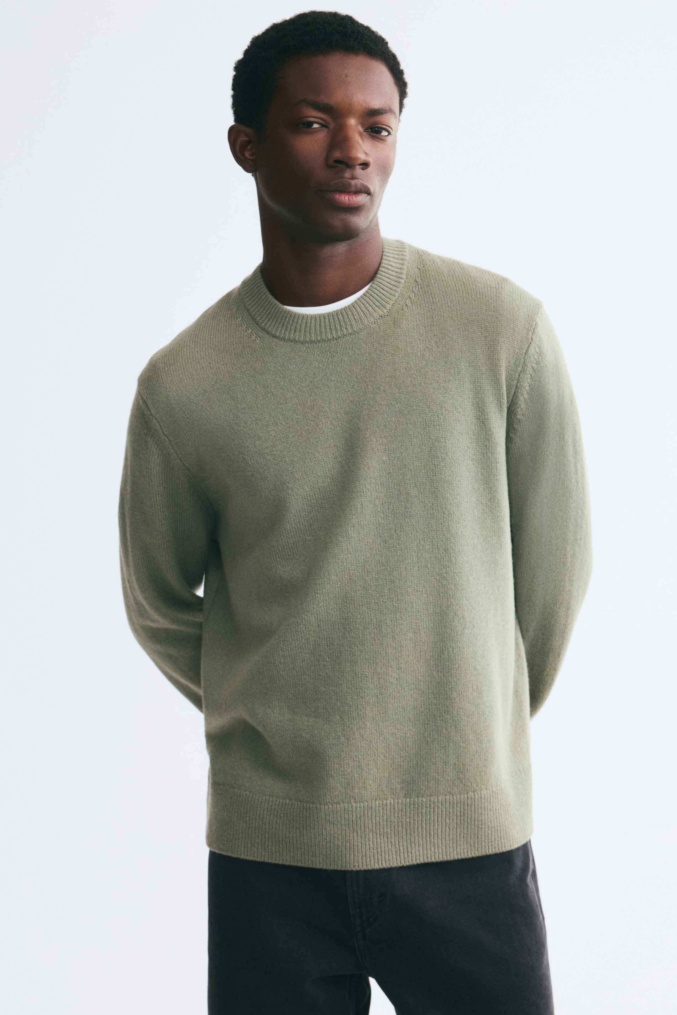 Größeres Bild anzeigen: Der Model trägt einen hellgrünen, gestrickten Wollpullover mit rundem, geripptem Ausschnitt und geripptem Saum. Der Pullover wird über einem weißen T-Shirt getragen und mit einer schwarzen Hose kombiniert.