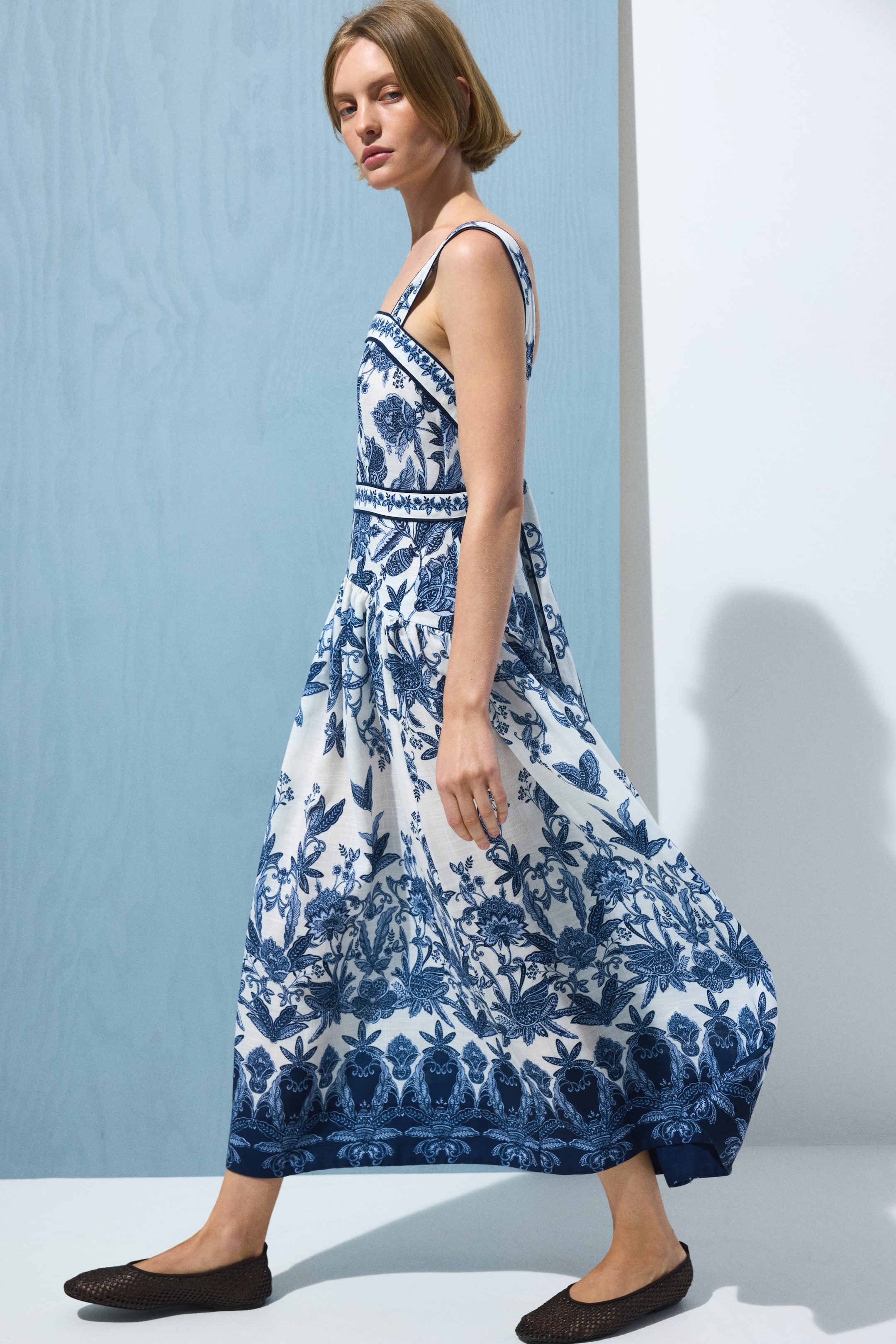 View larger image: The model is wearing a calf-length dress crafted from white woven fabric adorned with a blue botanical print. As she walks, the gently flared skirt billows slightly. The dress boasts a straight neckline, wide shoulder straps that cross at the back, and a fitted bodice with shaping seams. A gathered seam at the hips adds a touch of elegance. Complementing the look, the model is also wearing dark flat shoes.