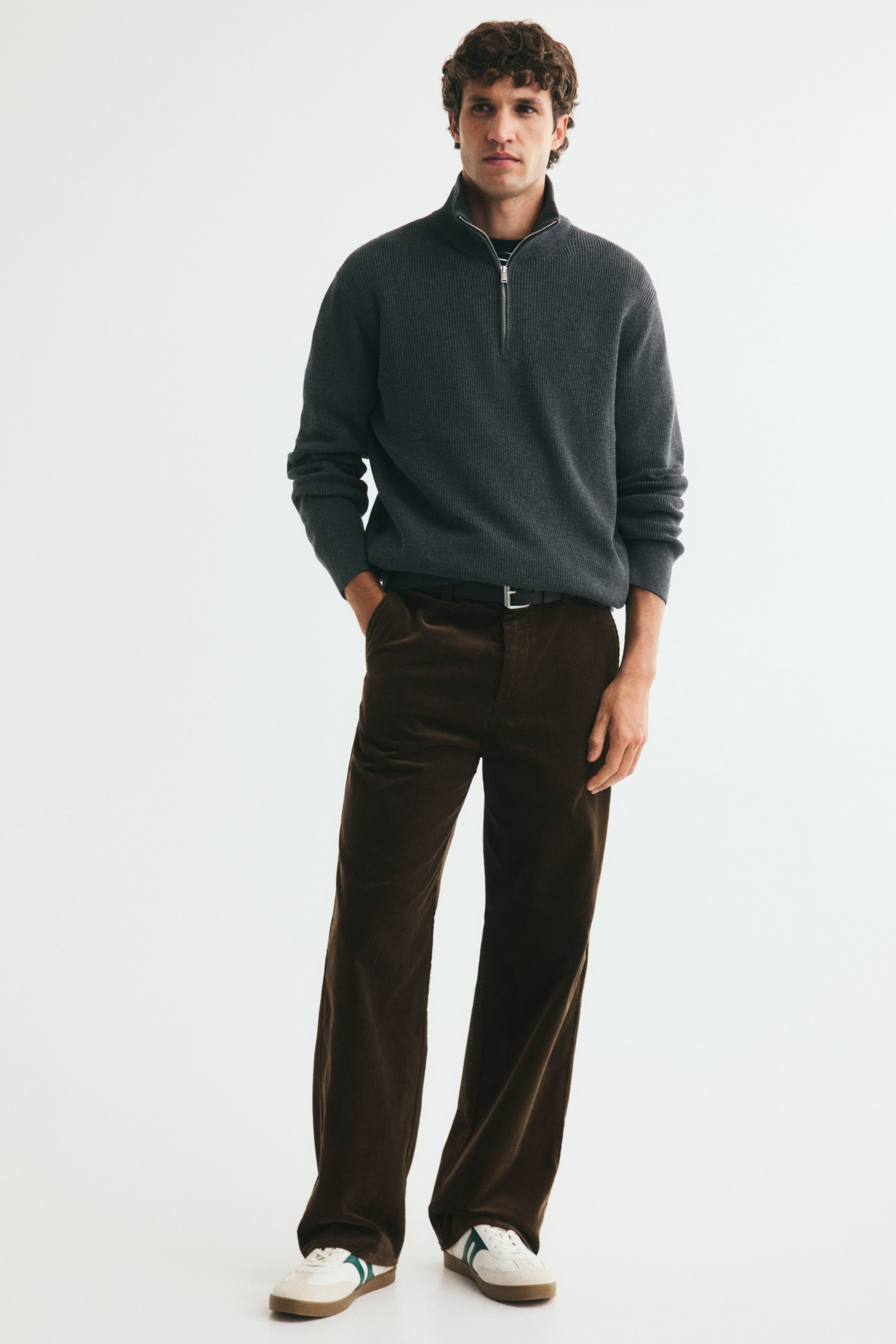Voir une image plus grande: Le mannequin porte un pantalon en velours côtelé brun foncé à coupe décontractée, en position debout, avec une main reposant dans une poche latérale diagonale. Le pantalon est maintenu par une ceinture foncée à la taille et est porté avec un chandail en tricot gris à demi-fermeture éclair et des espadrilles blanches.