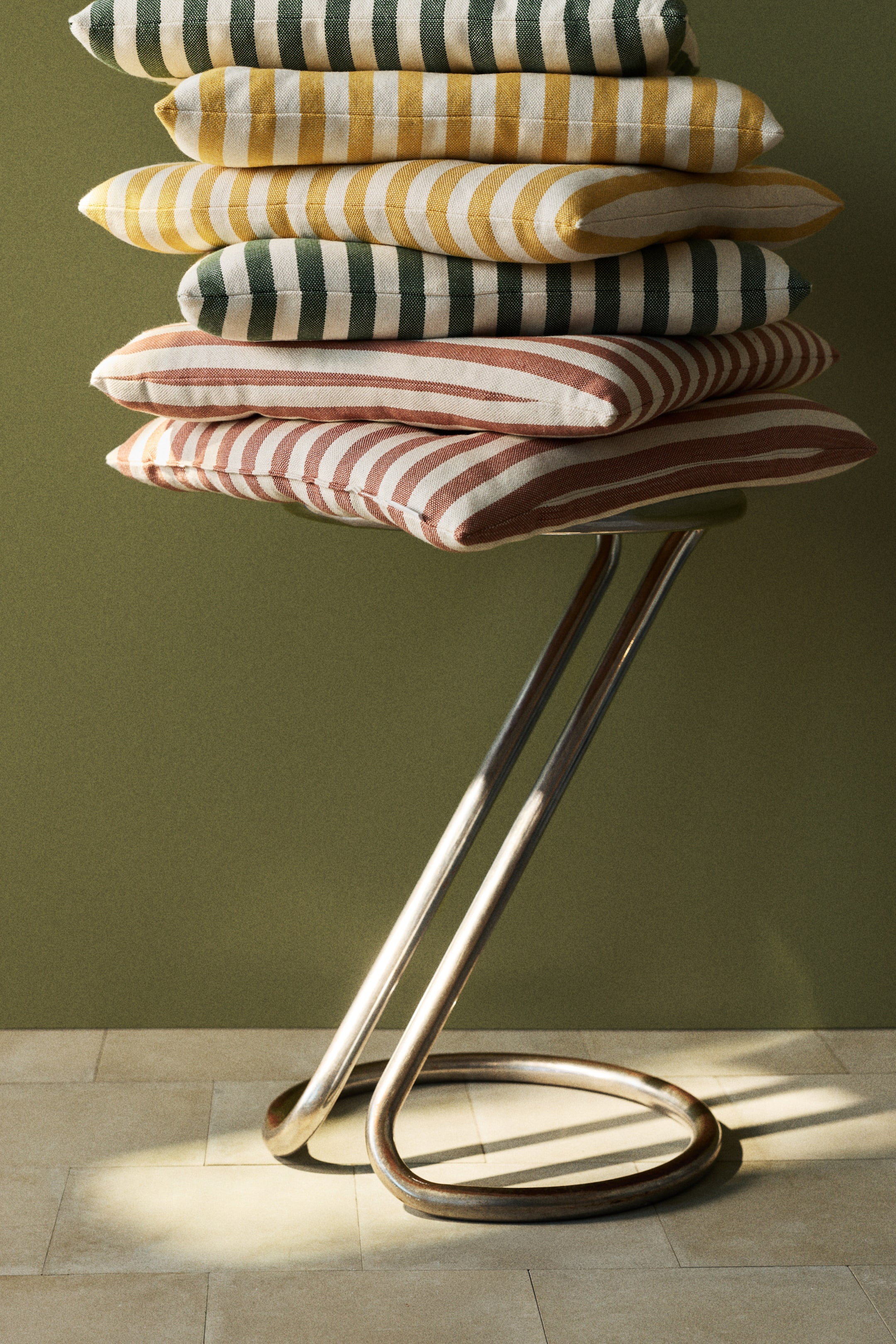 Vezi imaginea mai mare: Pernele de scaun căptușite sunt stivuibile, fiecare având o textură țesută cu dungi orizontale în culori alb-gălbui și verde închis, alb-gălbui și galben, și alb-gălbui și maro-roșiatic. Nodurile vizibile ale matlasării apar pe suprafața fiecărei perne, indicând construcția sa căptușită.