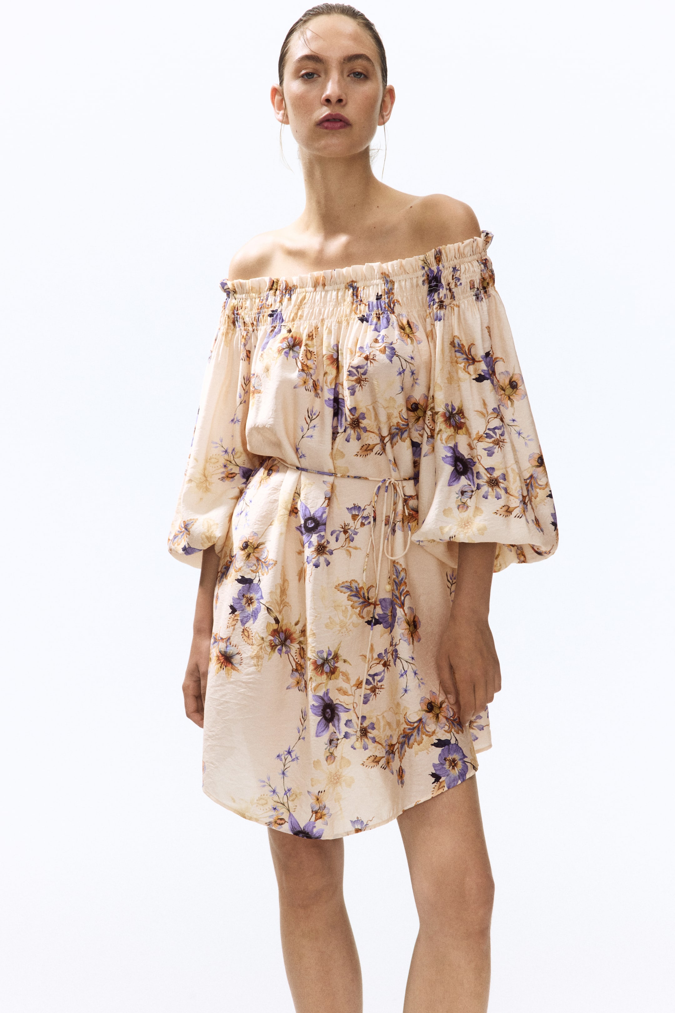 Ampliar la imagen: La modelo lleva puesto un vestido mini con estampado floral en color beige claro, con diseño de hombros descubiertos. El vestido cuenta con un escote fruncido, mangas voluminosas de tres cuartos y una cintura ajustable con cordones.