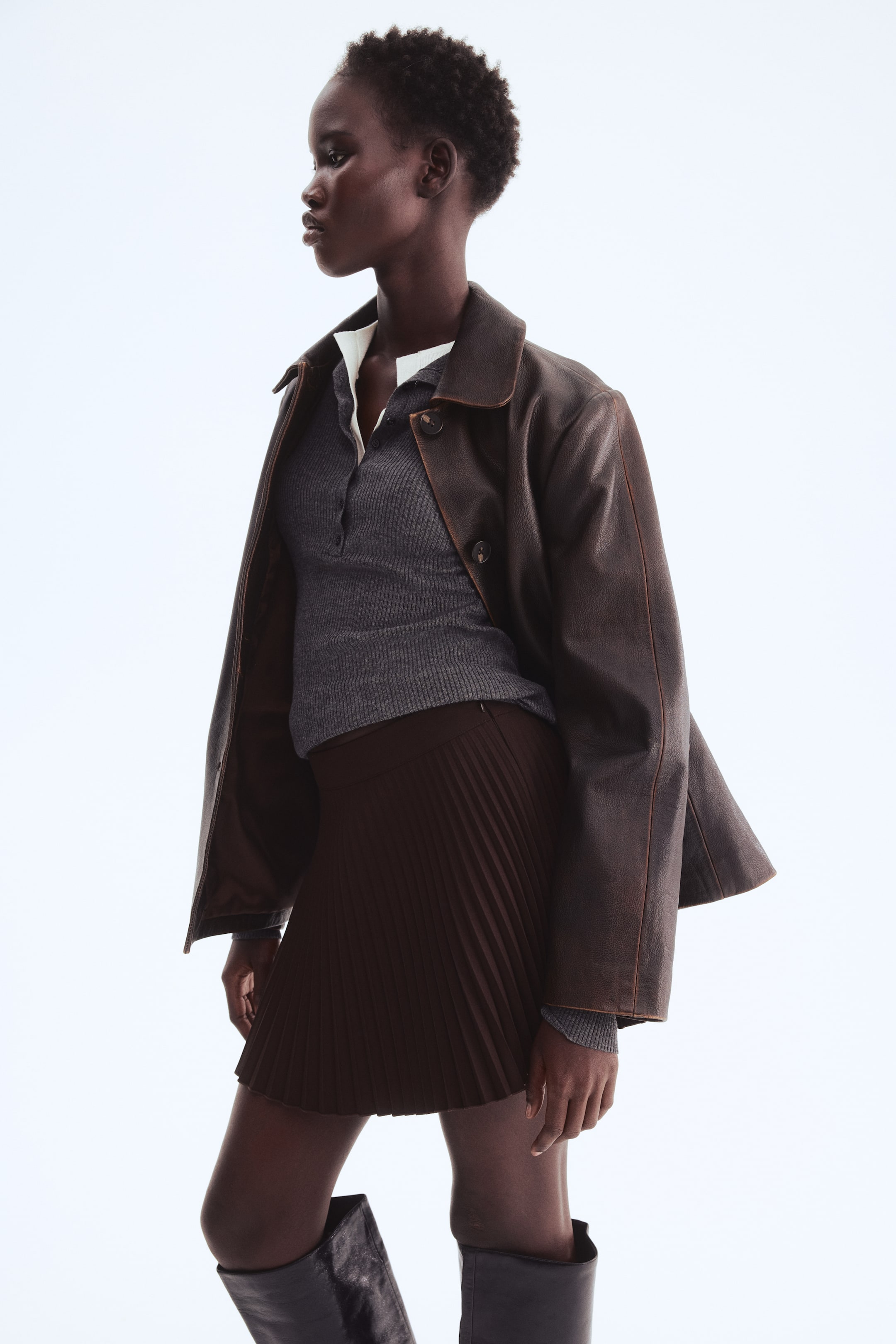 View larger image: A person is donning a full ensemble, posed at a slight angle to the right. They're sporting a dark brown, relaxed-fit collared jacket over a grey ribbed knit polo shirt, layered with a white collared top, and a dark brown pleated mini skirt paired with black knee-high boots.