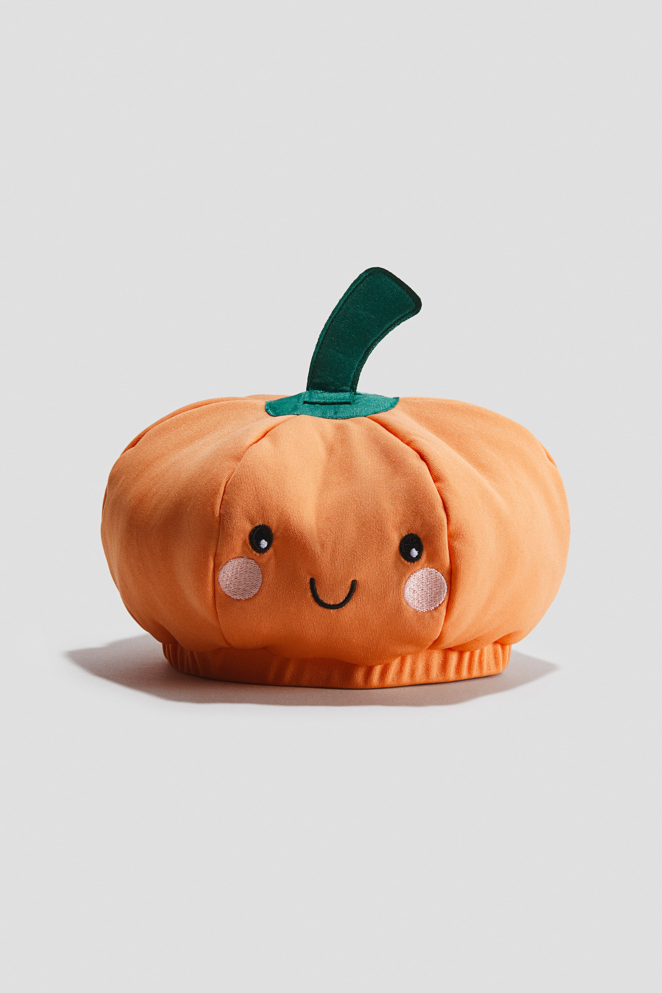 Voir une image plus grande: Un chapeau rembourré d'Halloween se présente sous la forme d'une citrouille orange, orienté vers l'avant, avec une tige verte sur le dessus, des yeux noirs brodés, une bouche incurvée et des marques de fard à joues roses en forme de cercles sur le devant, ainsi qu'une base élastiquée froncée.