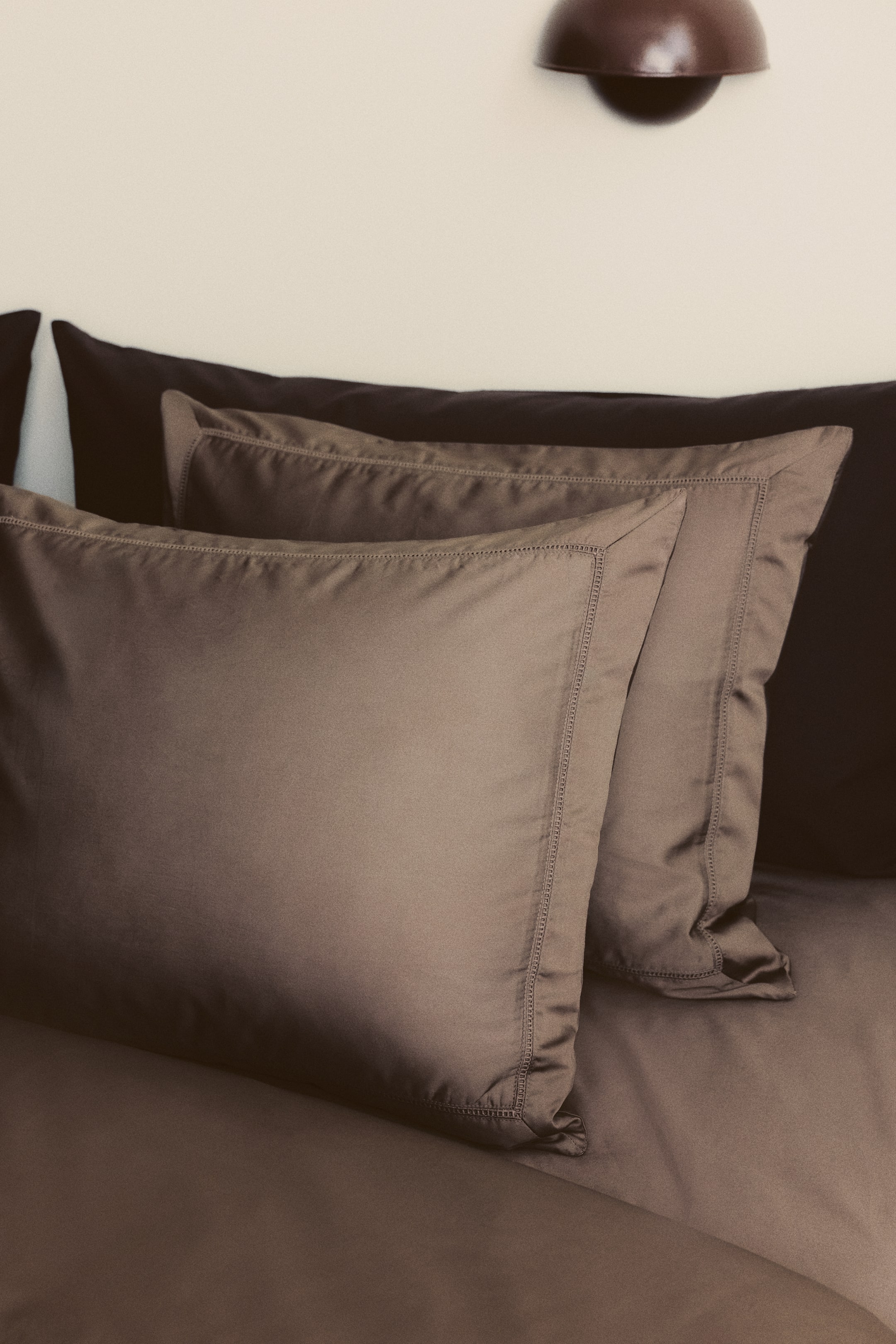 Agrandir l'image: Deux coussins taupe, ornés de bordures décoratives cousues autour de leurs bords, reposent légèrement inclinés sur un coussin brun foncé et une couette assortie.