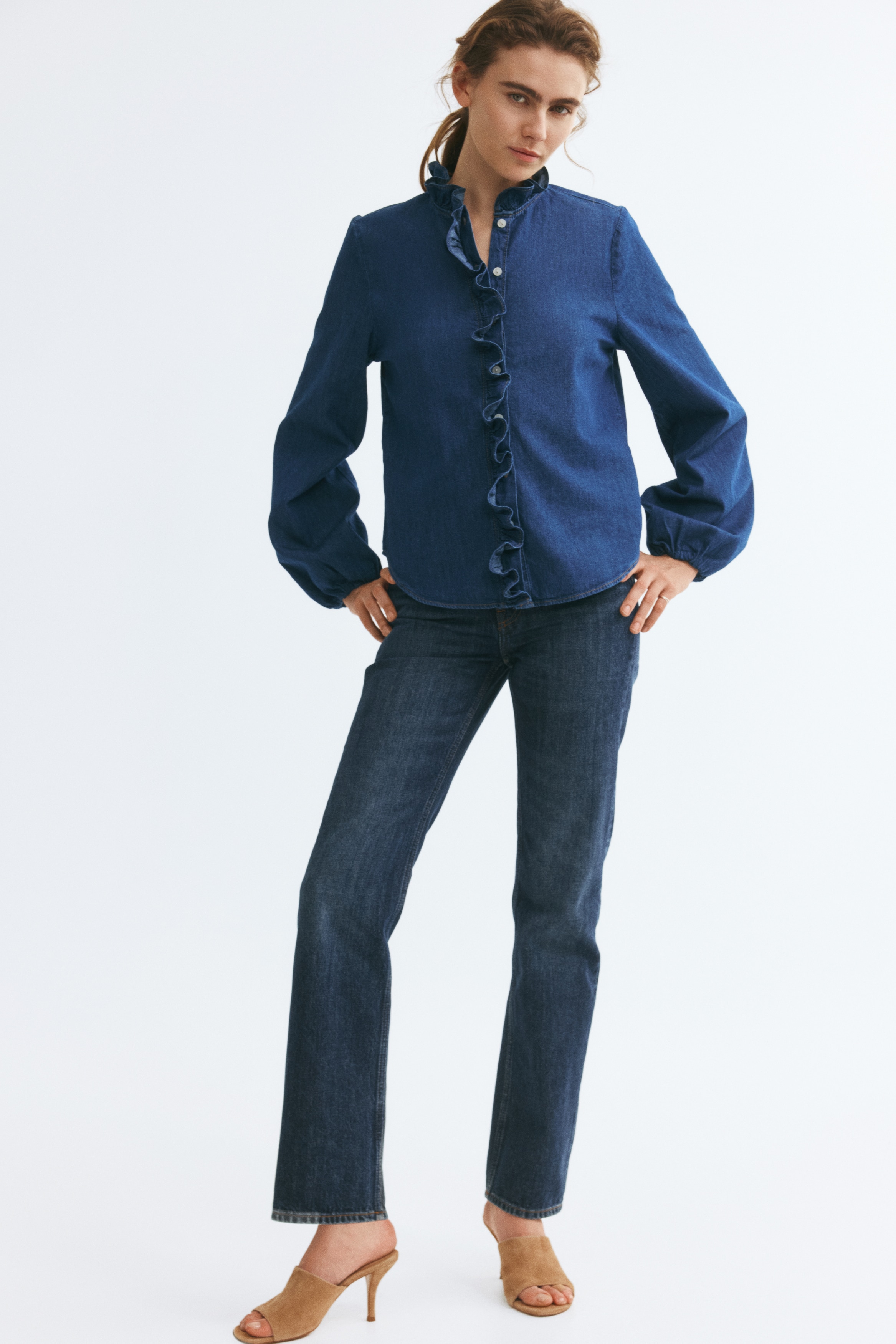 A blue denim blouse is hanging facing forwards, showing a ruffled stand collar and matching ruffled trim along the centre front placket. The blouse features long, voluminous balloon sleeves with gathered cuffs and a curved hem.