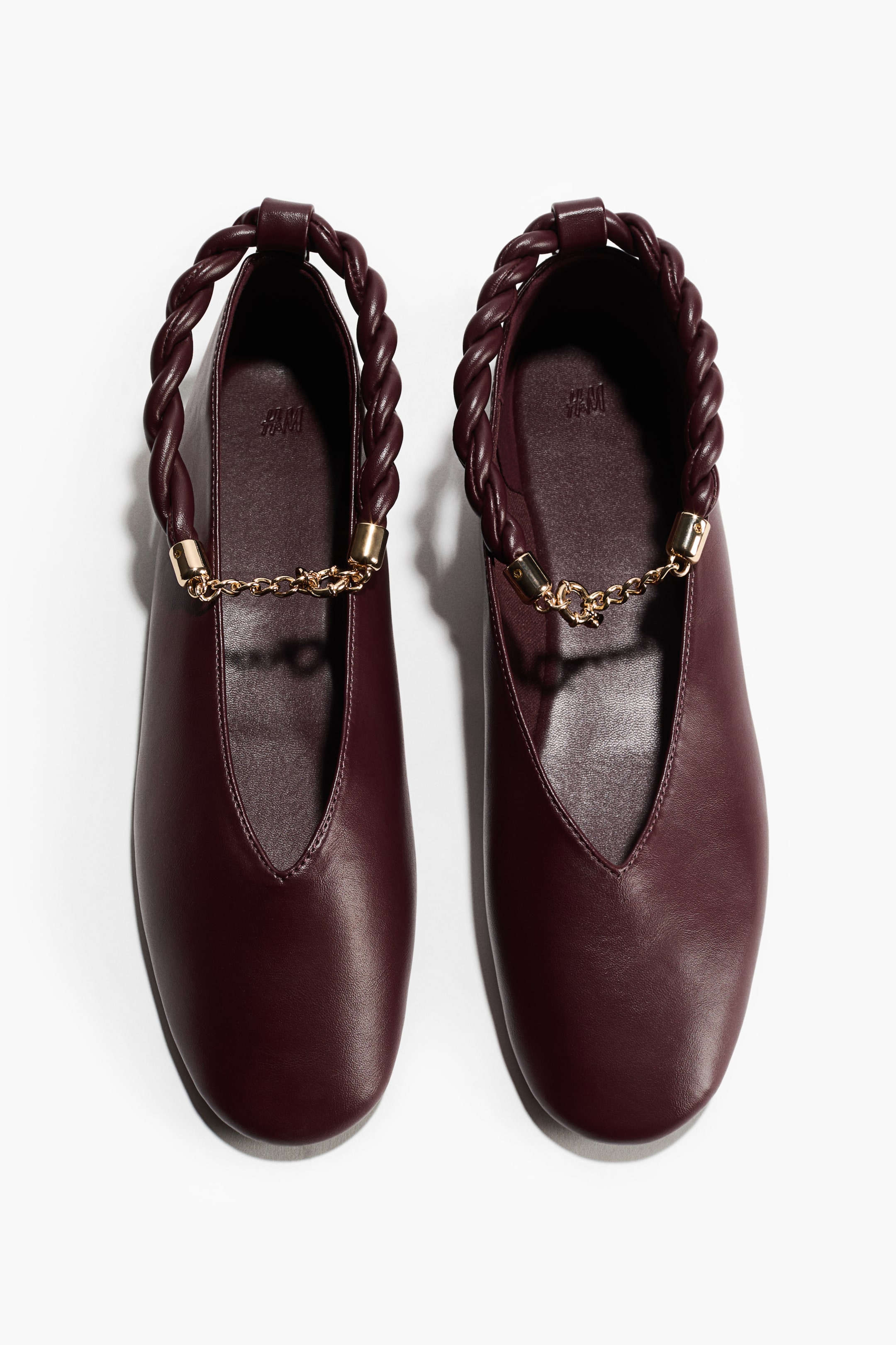 Agrandir l'image: Le produit est une paire de ballerines bordeaux, présentées de FACE, avec chaque chaussure dotée d'un bout arrondi et d'un décolleté en V. Une bride torsadée traverse l'ouverture supérieure de chaque chaussure, ses extrémités étant fixées par des anneaux dorés qui se connectent également à une délicate chaîne dorée.