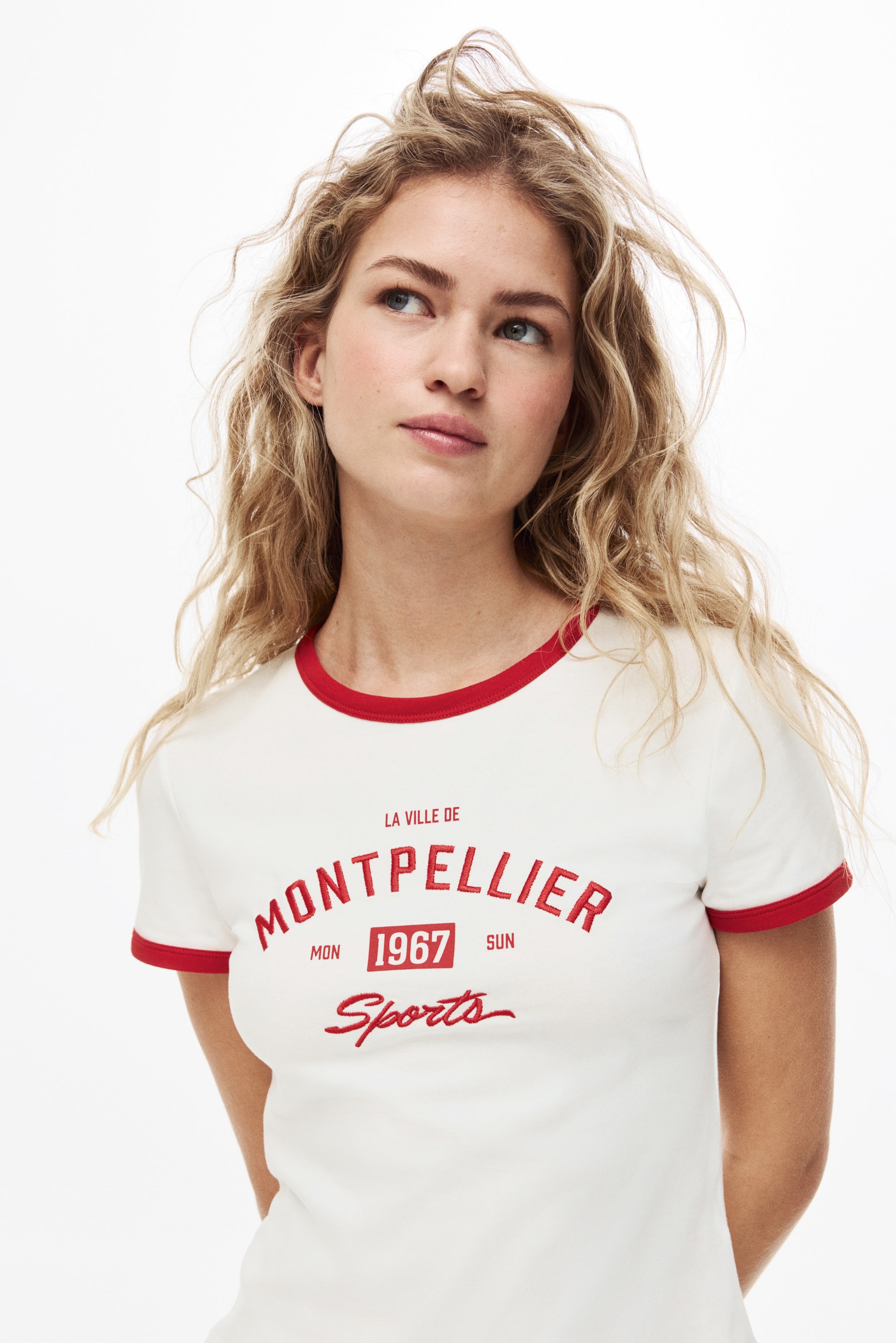 View larger image: An off-white ringer t-shirt features red trim on its crew neck and short sleeve cuffs, displayed at a slight angle. Red text is printed on the front, reading "LA VILLE DE MONTPELLIER 1967 MON SUN Sports."