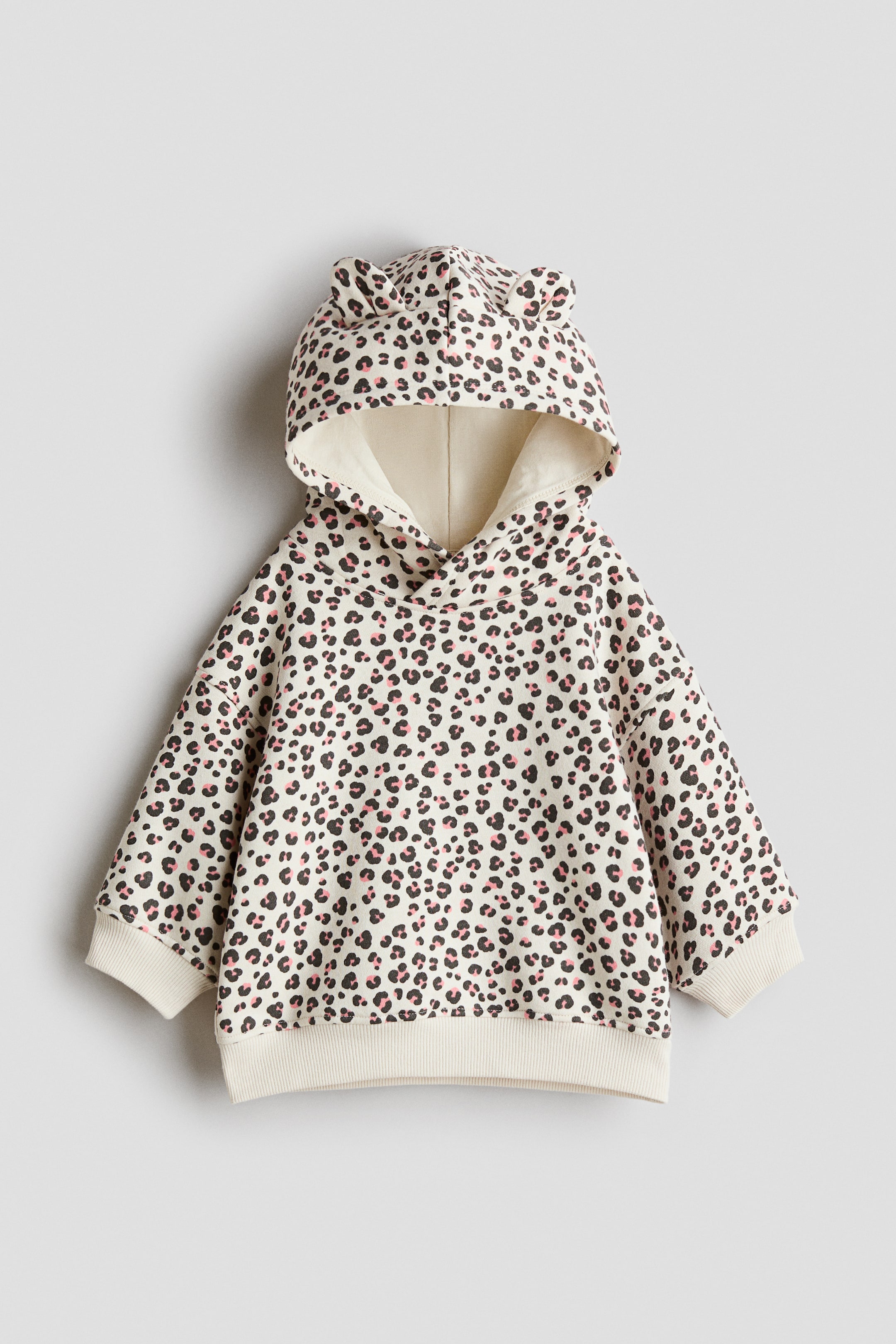 Voir une image plus grande: Vue de face : Un chandail à capuchon crème présente un imprimé léopard avec des contours noirs et des centres roses partout. Il présente une capuche avec de petites oreilles arrondies, des manches longues avec des poignets côtelés et un ourlet côtelé.