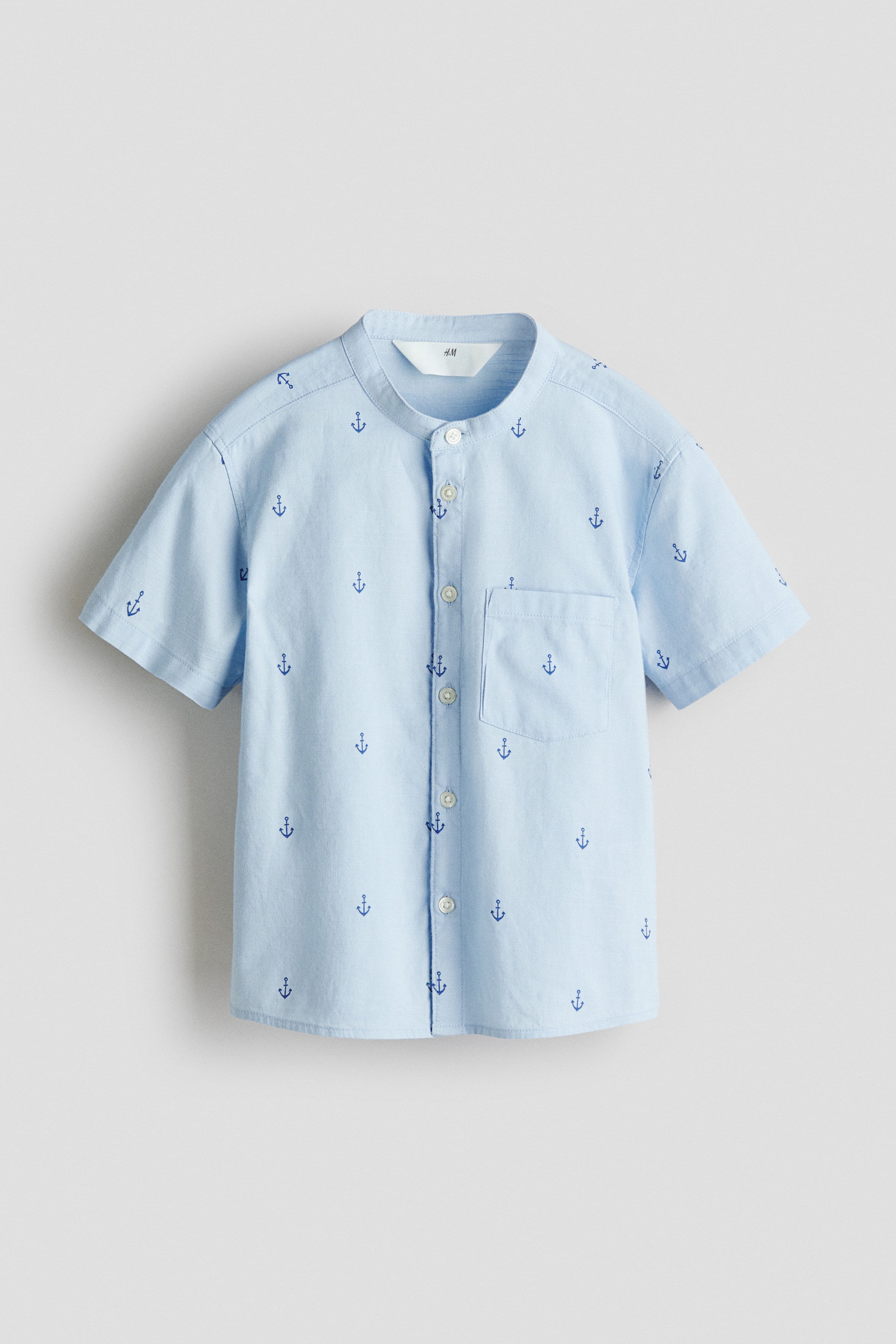 View larger image: FRONT of a loose-fit shirt, appearing in light blue and displaying a repeating pattern of small dark blue anchors. It features a band collar, buttons down the front, and has an open chest pocket on the left side, with short sleeves.