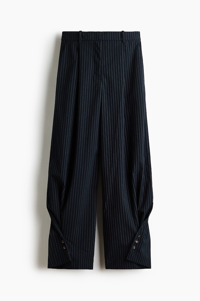 A pair of navy blue, pinstriped woven pants is hanging facing front, displaying a high waist with front pleats. The barrel-leg silhouette is tapering towards the bottom, showcasing unique wrap-around sides secured with buttons at the outer hems. - 1