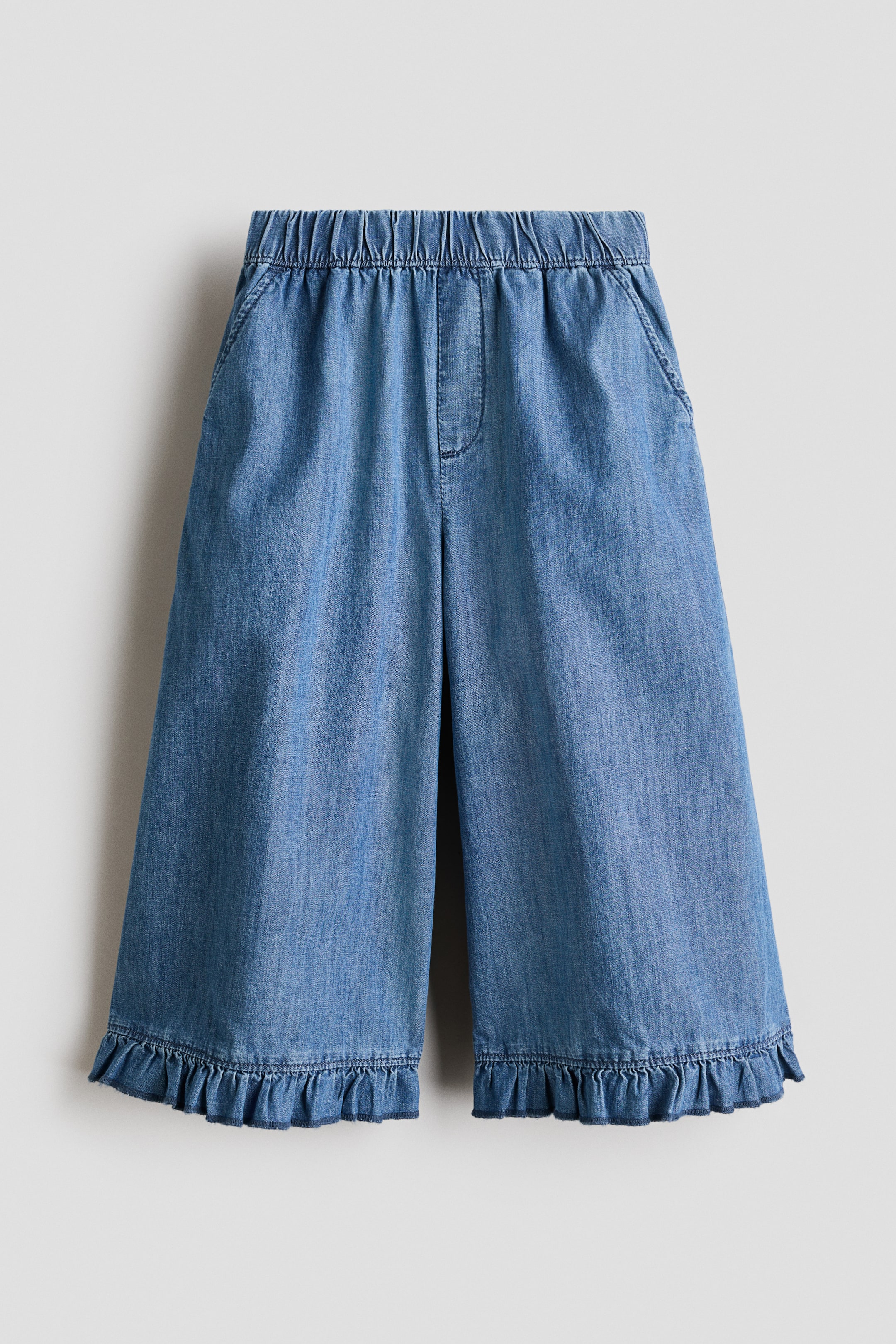 View larger image: Light blue denim culotte pants come with an elastic waistband and two side slash pockets. They showcase a wide-leg fit with ruffled hems at the bottom.