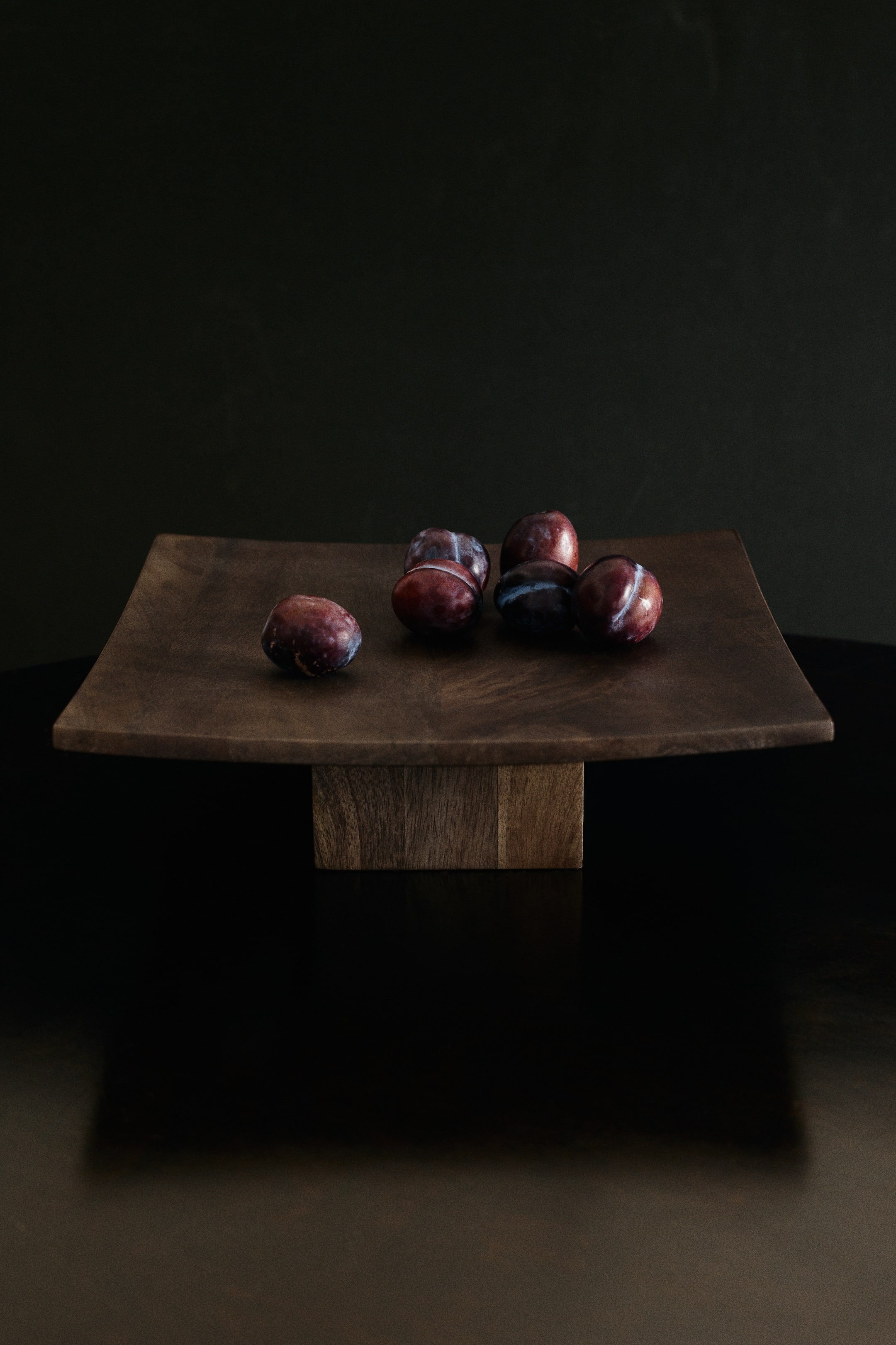 Größeres Bild anzeigen: Ein quadratisches Serviertablett mit Fuß aus dunkel getöntem Mangoholz ist von vorne zu sehen, mit einer leicht gewölbten quadratischen Platte, die von einem dicken, kantigen Sockel getragen wird. Sechs dunkelviolette Pflaumen liegen auf der Oberfläche des Tabletts und heben die sichtbare natürliche Holzmaserung hervor.