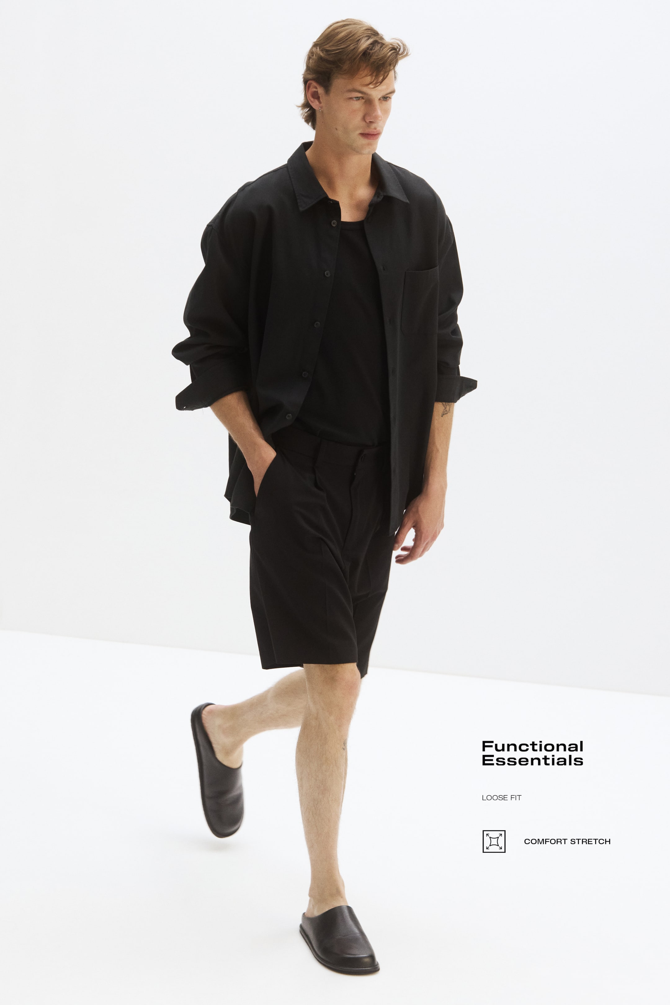 View larger image: A black loose-fit button-up shirt is showcased open over a black t-shirt, paired with black mid-thigh length shorts and black slip-on shoes. The shirt features a collar and a front patch pocket, with its sleeves casually rolled up, all viewed from a slightly angled front perspective.