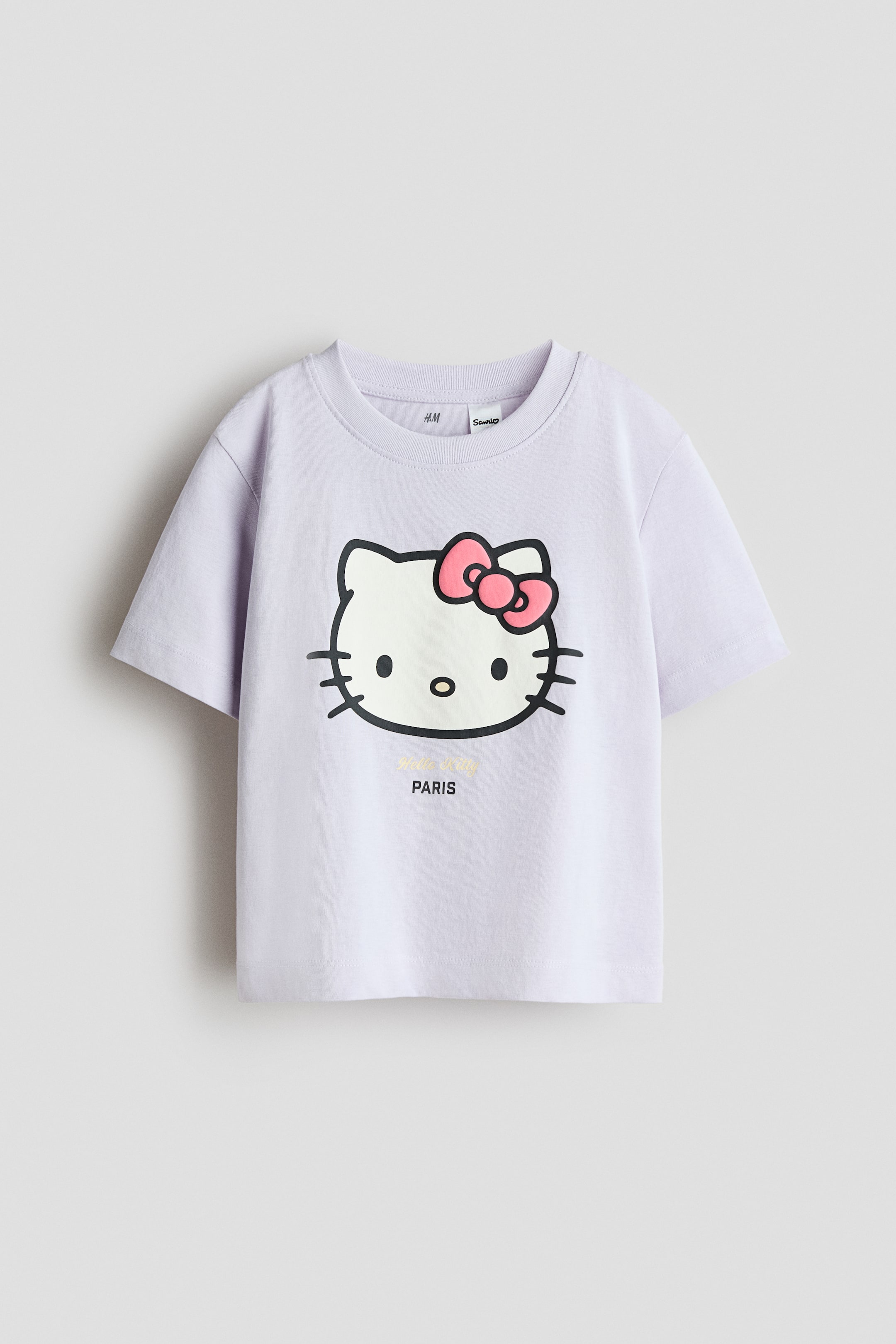 View larger image: A light purple T-shirt is showcasing its front view, crafted from soft cotton jersey, featuring a round, ribbed neckline and gently dropped shoulders. A large graphic of Hello Kitty's head with a pink bow is printed centrally on the front, with 'Hello Kitty' in gold script and 'PARIS' in black letters positioned below.
