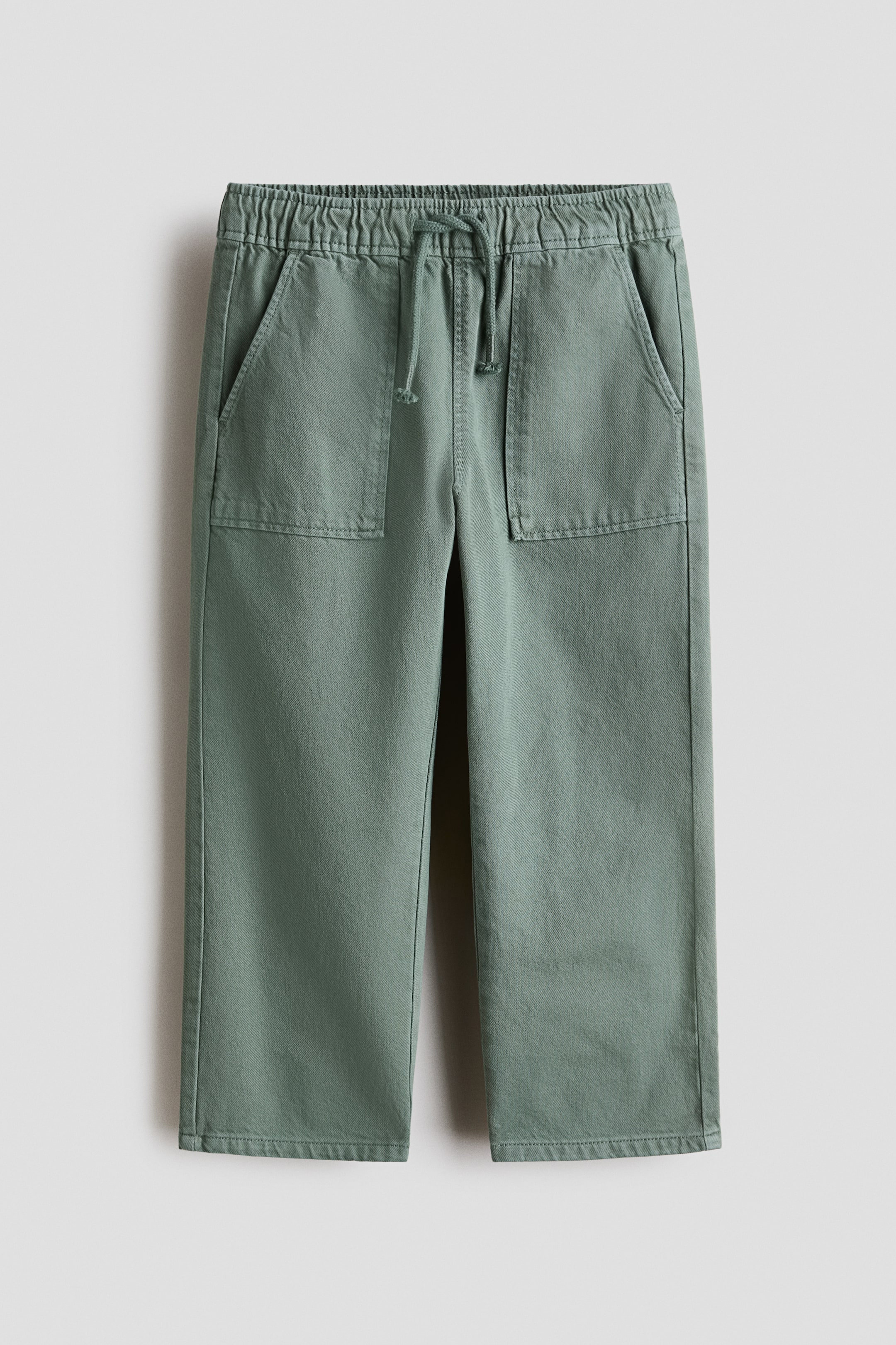 View larger image: Green trousers are presenting a front view, featuring an elasticated waistband with a drawstring fastening and two side slash pockets. Two large rectangular patch pockets are positioned on the upper front legs, and the trousers are designed with a relaxed, straight fit ending in a stitched hem.
