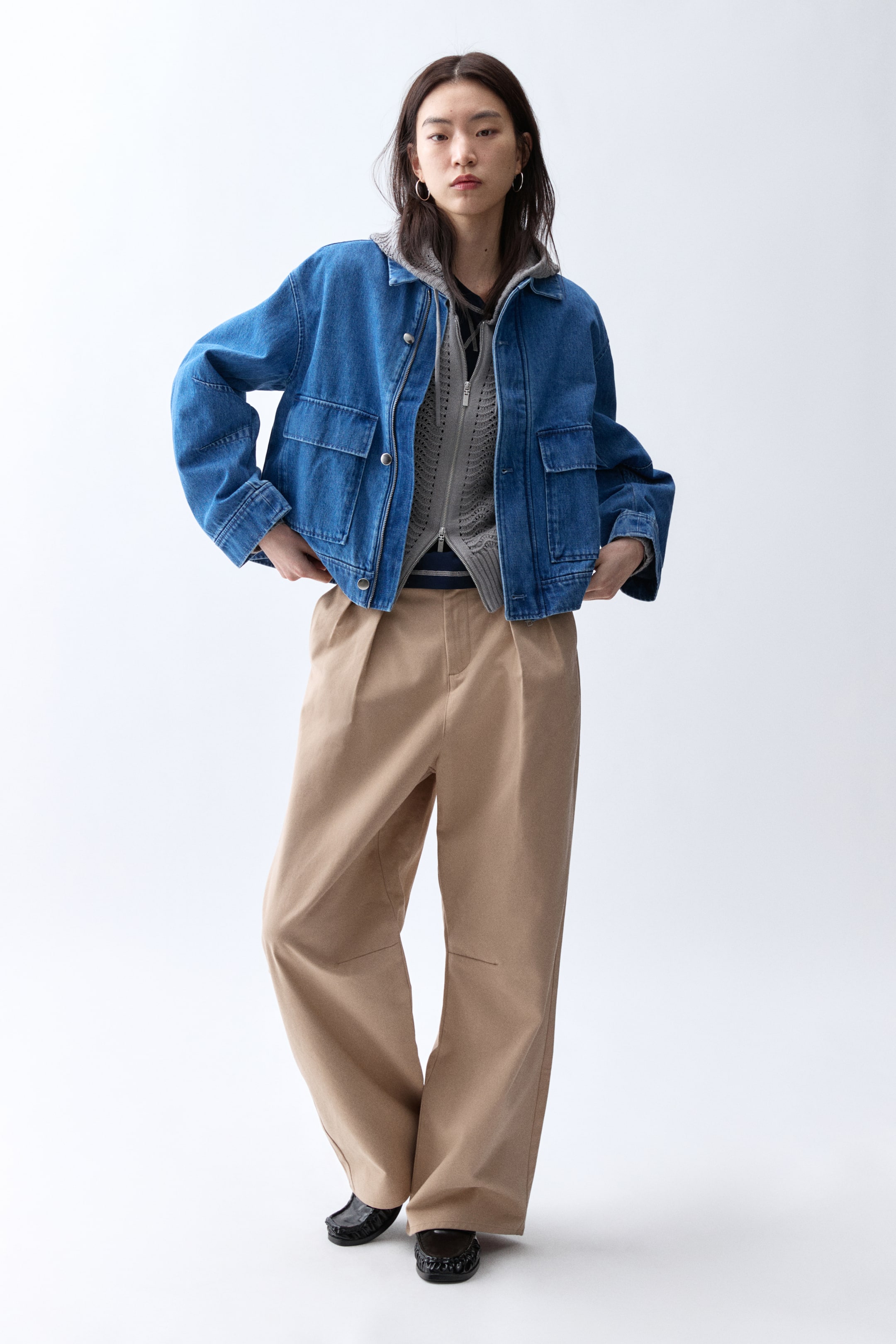 Xem ảnh lớn hơn: Một chiếc áo khoác denim xanh lam có kiểu dáng rộng rãi, thoải mái với cổ áo tiêu chuẩn và hàng nút cài phía trước màu bạc, nhìn từ góc phía trước. Nó có hai túi nắp lớn ở ngực và hai túi vá phía dưới, được xếp lớp bên ngoài áo hoodie xám và áo cardigan len xám có khóa kéo.