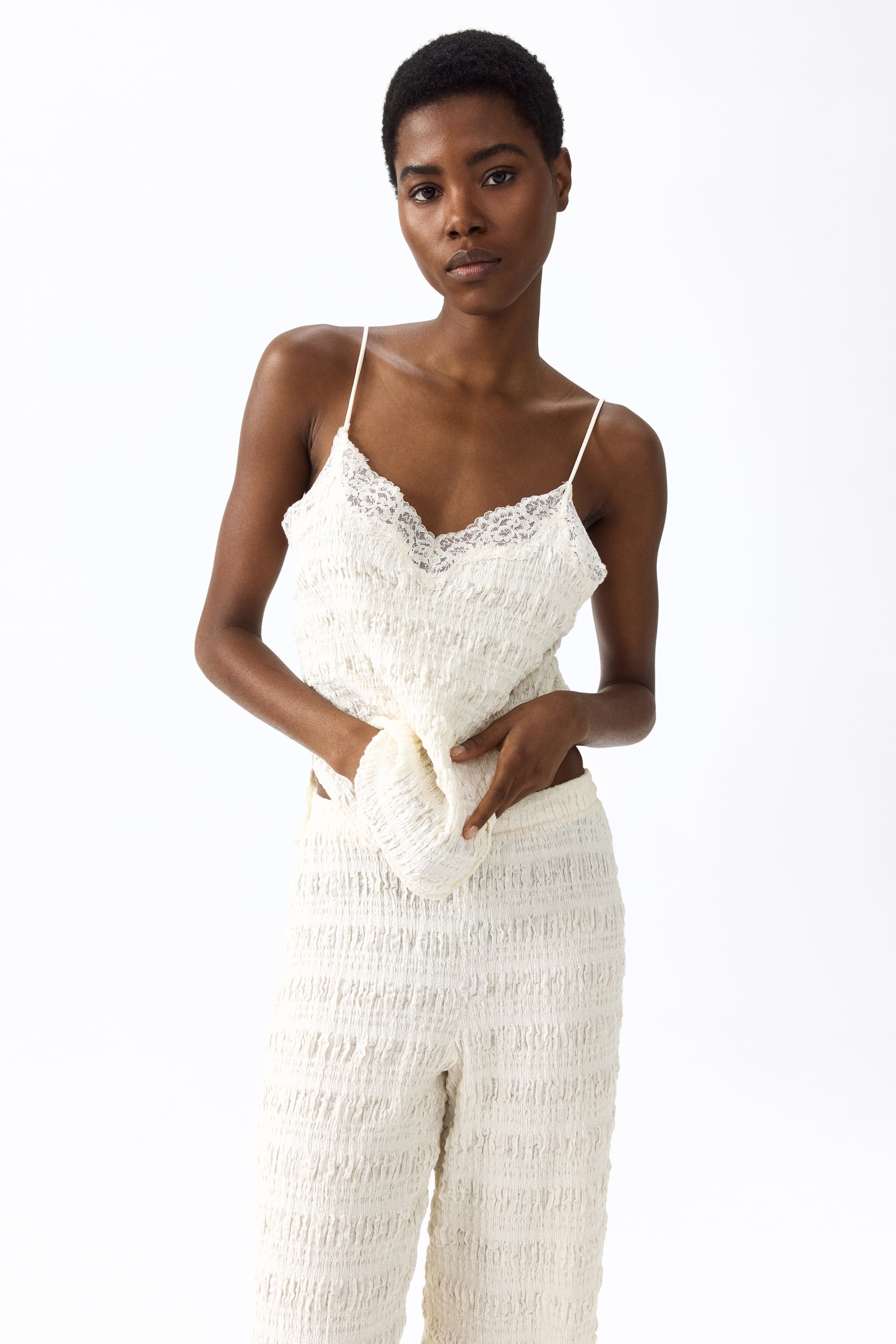 Visualizza immagine più grande: Un completo a due pezzi color crema viene presentato, caratterizzato da un top con spalline sottili e bordatura in pizzo bianco che rifinisce lo scollo a V, e pantaloni a gamba larga abbinati con effetto arricciato. Il set è posizionato con un angolo leggermente frontale a sinistra, con il top delicatamente tirato in avanti per mettere in risalto il tessuto testurizzato e la vestibilità rilassata.