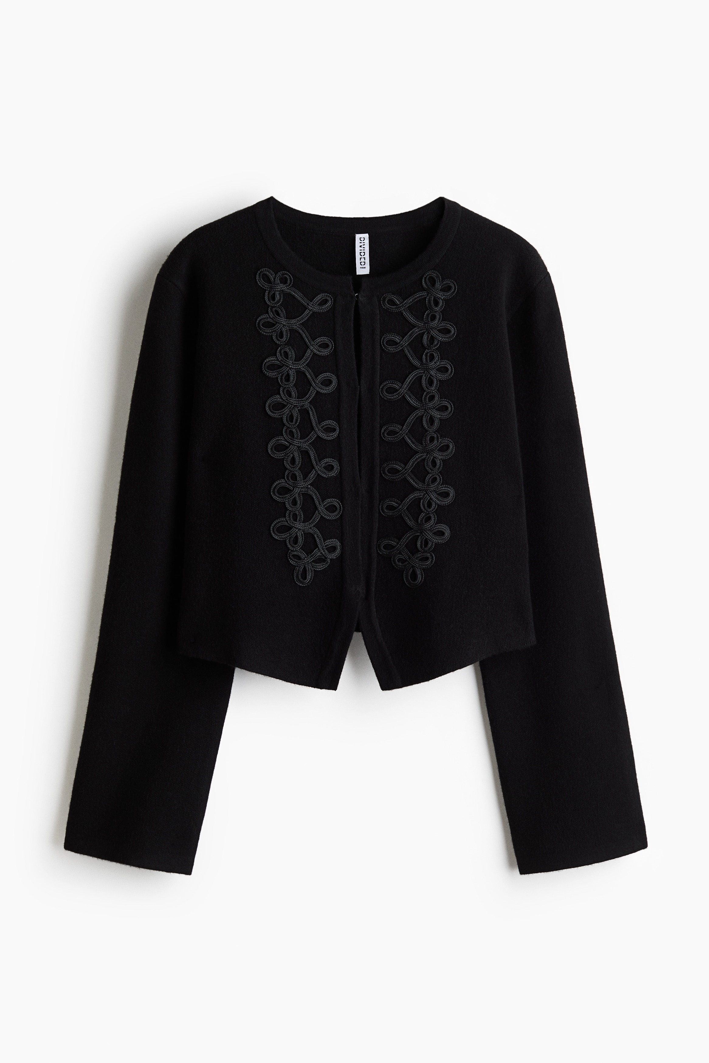 The black Short Rope Detail Cardigan is worn by the model, featuring a crew neckline, long sleeves, and symmetrical rope embroidery along the front placket. The cardigan is styled with white trousers as the model stands with crossed arms.