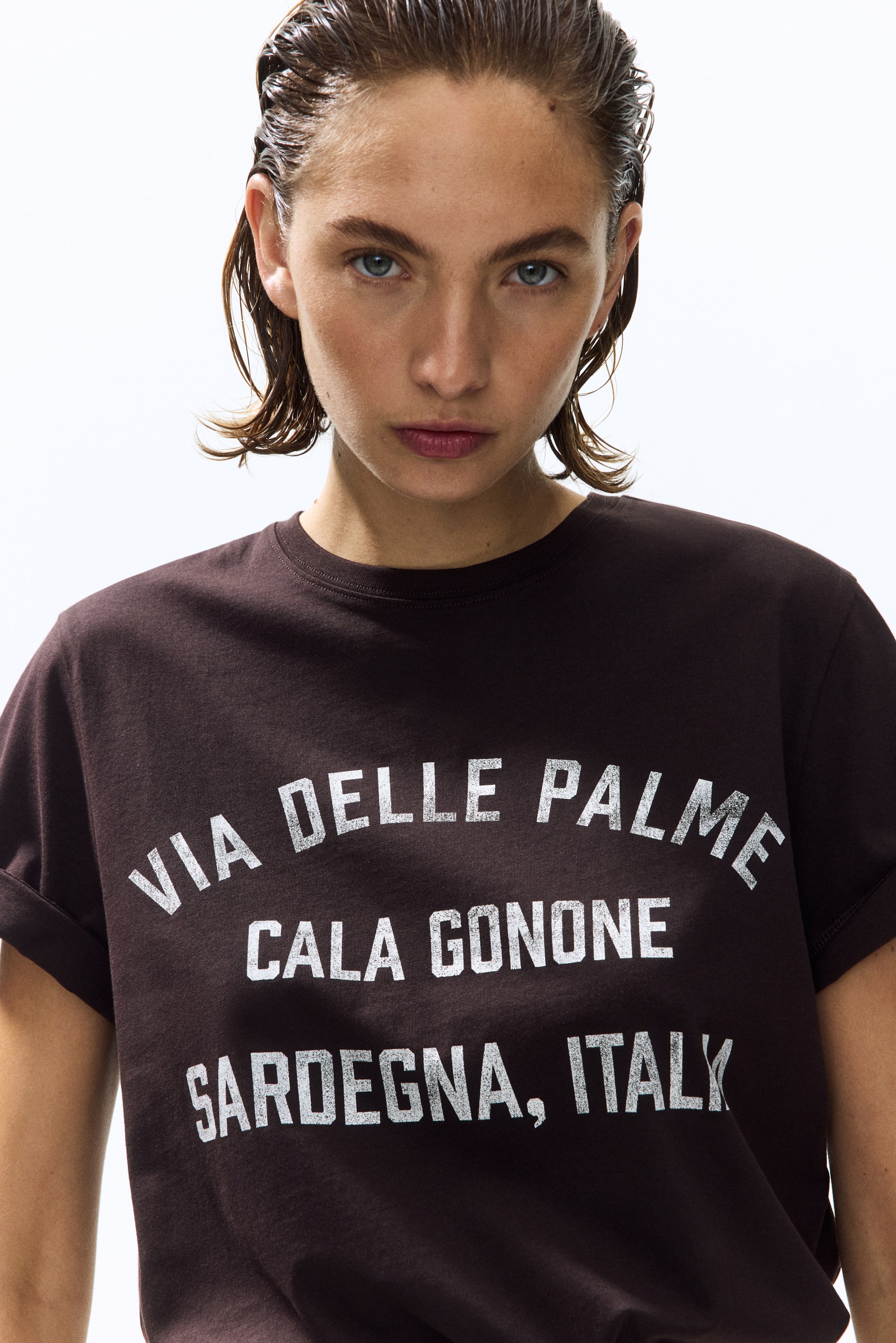 View larger image: The model is wearing a dark brown cotton jersey t-shirt with a round neckline. It showcases a white distressed text print on the front that reads "VIA DELLE PALME CALA GONONE SARDEGNA, ITALY," and features cuffed short sleeves.