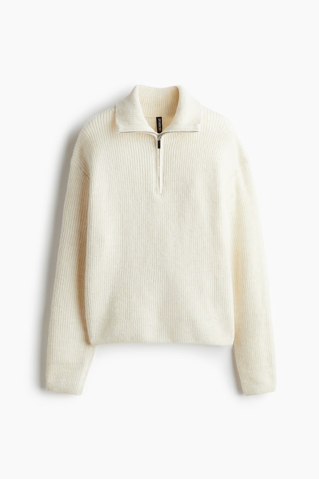 A cream-colored knitted sweater is being presented from the front, featuring a quarter-zip closure with a silver-tone zipper and a standing collar. The sweater's body and long sleeves are showcasing a vertical ribbed knit. - 1