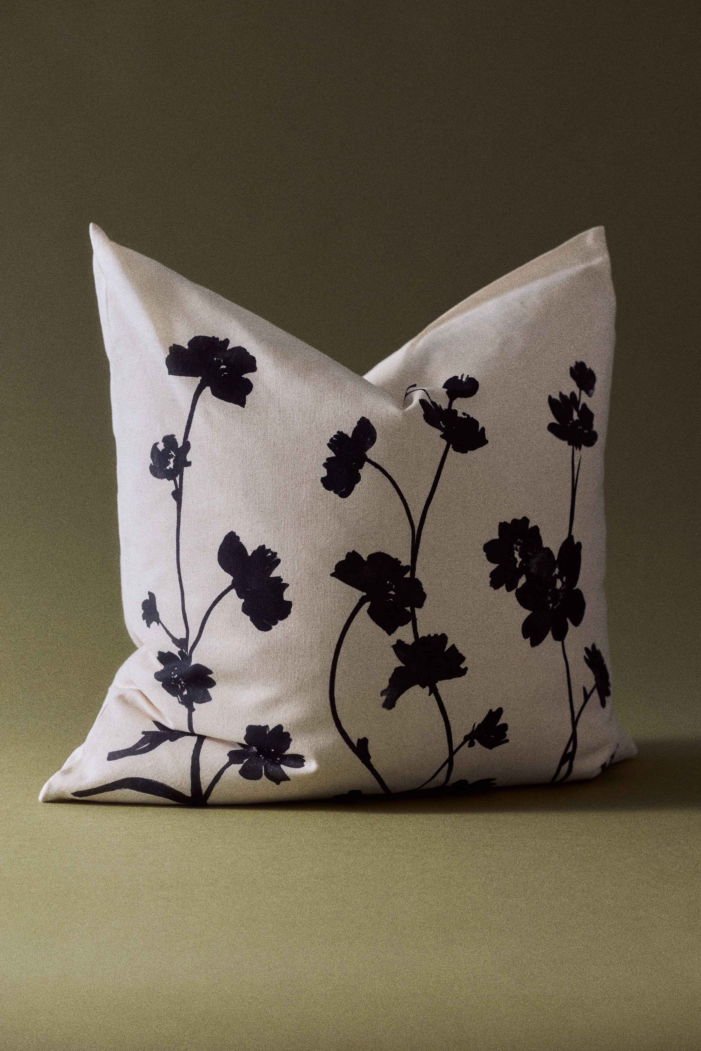 Een kussenhoes van katoenen canvas toont zijn voorkant, met een botanische print van zwarte gestileerde bloemen en stelen op een gebroken witte achtergrond.