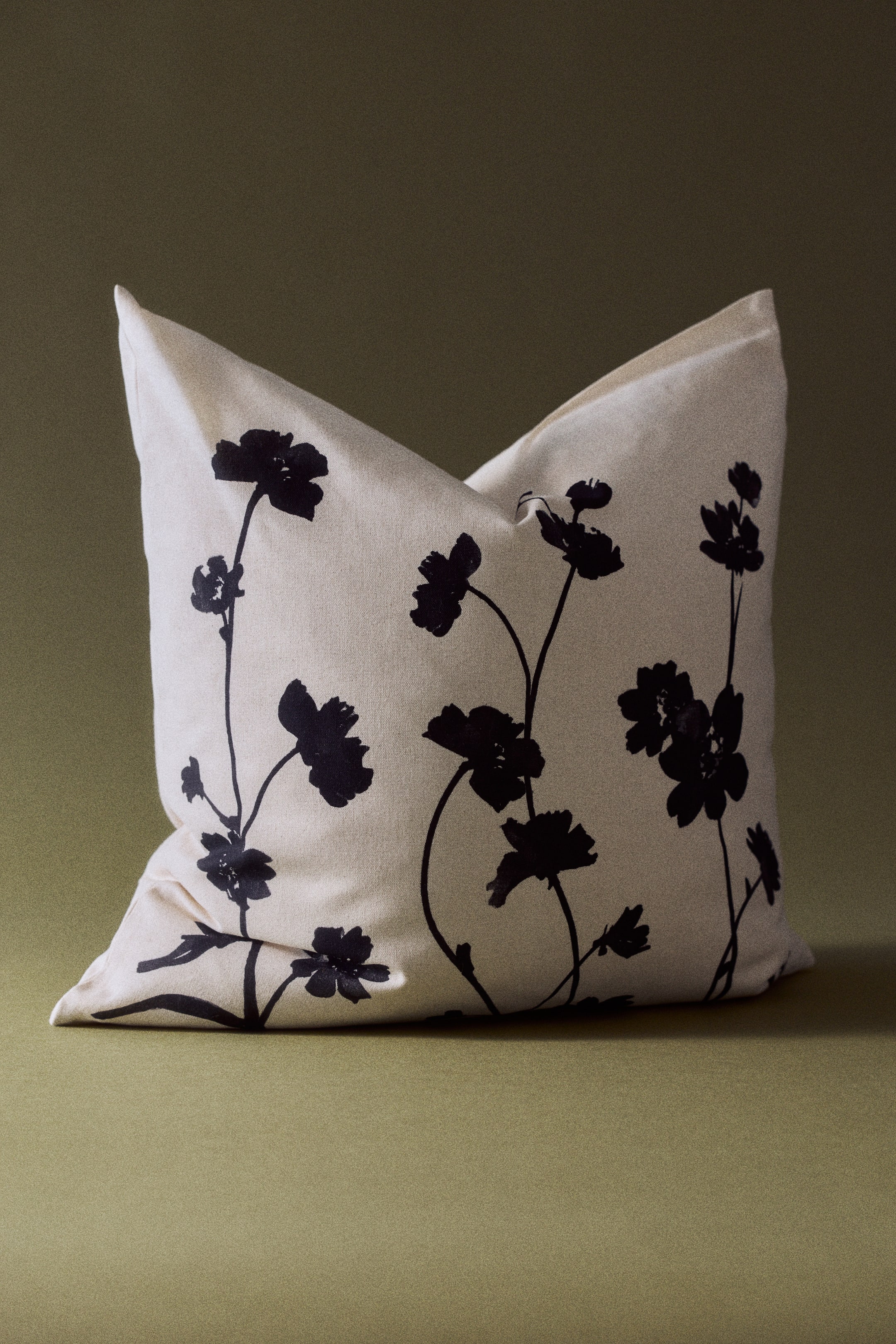 View larger image: A square cotton canvas cushion cover in light cream stands upright and faces forward, showcasing a black botanical print of wildflower silhouettes on the front. The cushion has a soft indentation at the top, highlighting its woven canvas texture and structure.
