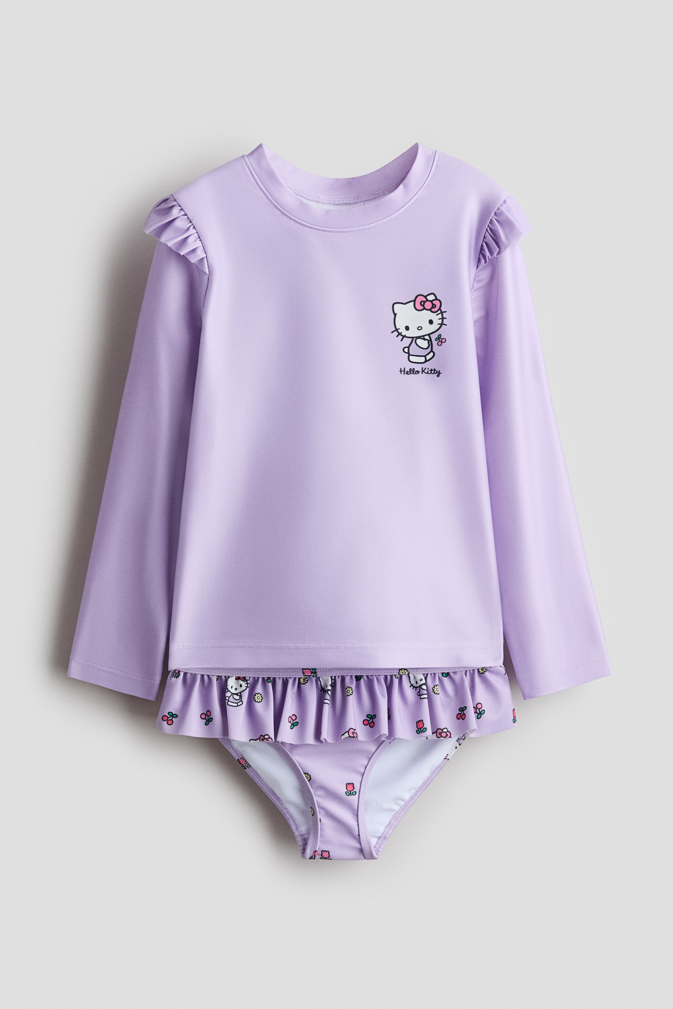 Visa större bild: Ett tryckt UV-skyddande badset med UPF 50 visar upp en lavendelfärgad långärmad topp och matchande bikinibyxor, båda vända framåt. Toppen har en Hello Kitty-grafik på framsidan, volangkanter på axlarna och en liten ståkrage, medan byxorna visar en volangkant i midjan och ett mönster av Hello Kitty-ansikten och små blommor.