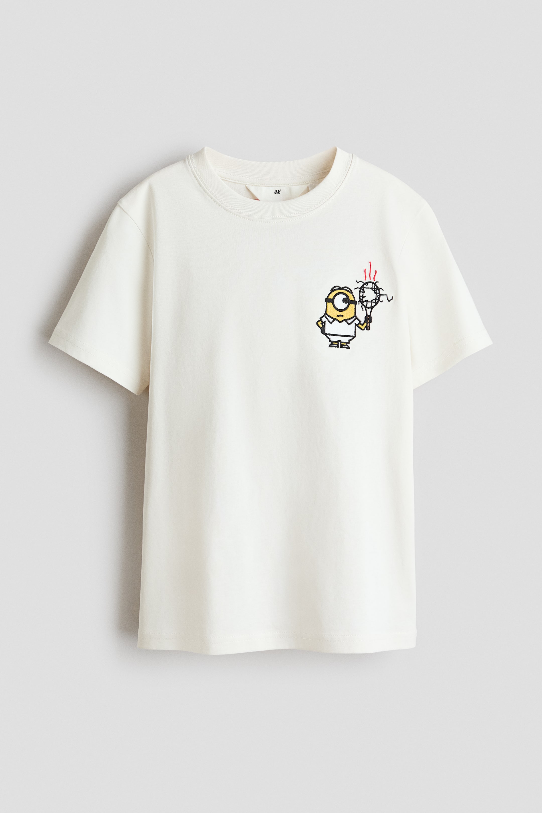 Ver imagem maior: Uma t-shirt em algodão jersey macio, de cor branco sujo, apresenta uma gola redonda com acabamento em malha canelada e ombros descaídos, com um gráfico de um personagem Minion amarelo segurando uma raquete de ténis com linhas vermelhas a aparecer no lado esquerdo do peito.