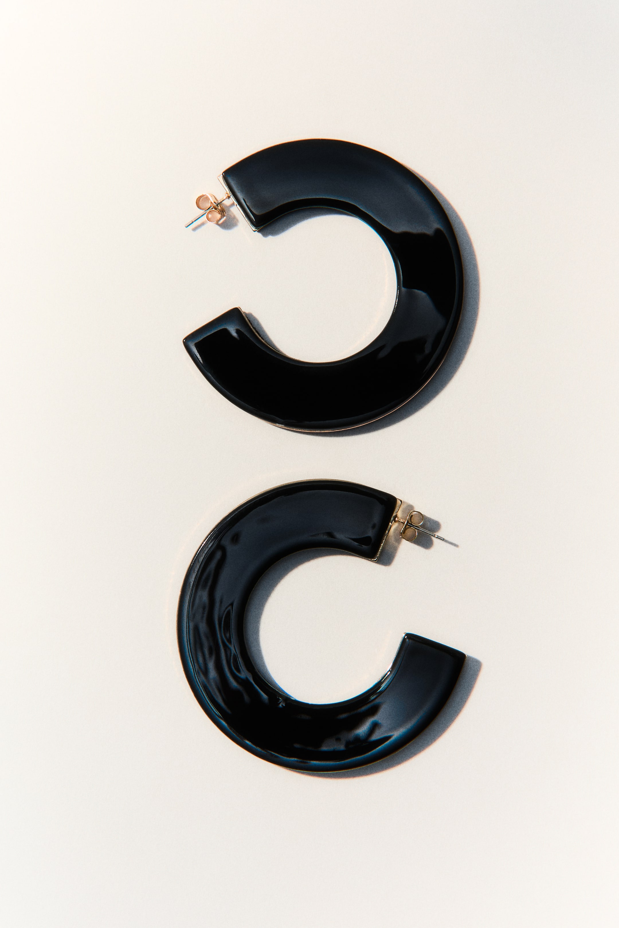 View larger image: Two black C-shaped earrings are lying flat, showcasing their reflective surfaces. The upper earring boasts a smooth finish, while the lower earring features a slightly wavy texture, with gold-toned posts visible on both.