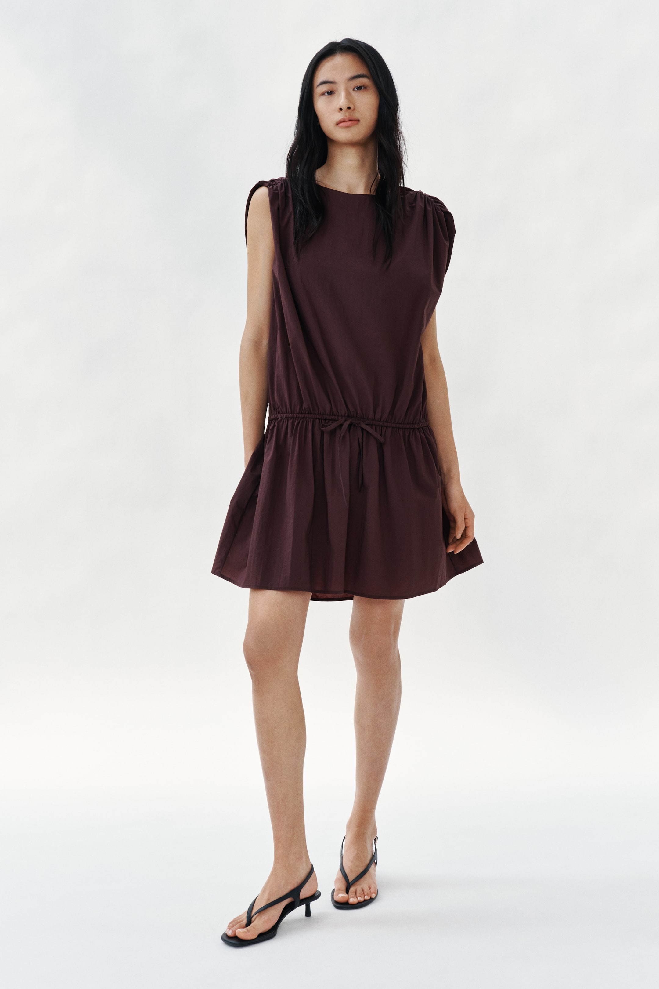 View larger image: Ang modelo ay nakasuot ng isang damit na hanggang tuhod, walang manggas, at gawa sa kayumangging hinabing tela. Ang damit ay may boat neckline, may mga gathered seam sa balikat para sa draped na epekto, malalapad na butas ng braso, at may drawstring sa ibaba ng balakang na lumilikha ng dropped-waist na epekto na may banayad na flare sa laylayan. Ang modelo ay nakasuot din ng itim na sandals.