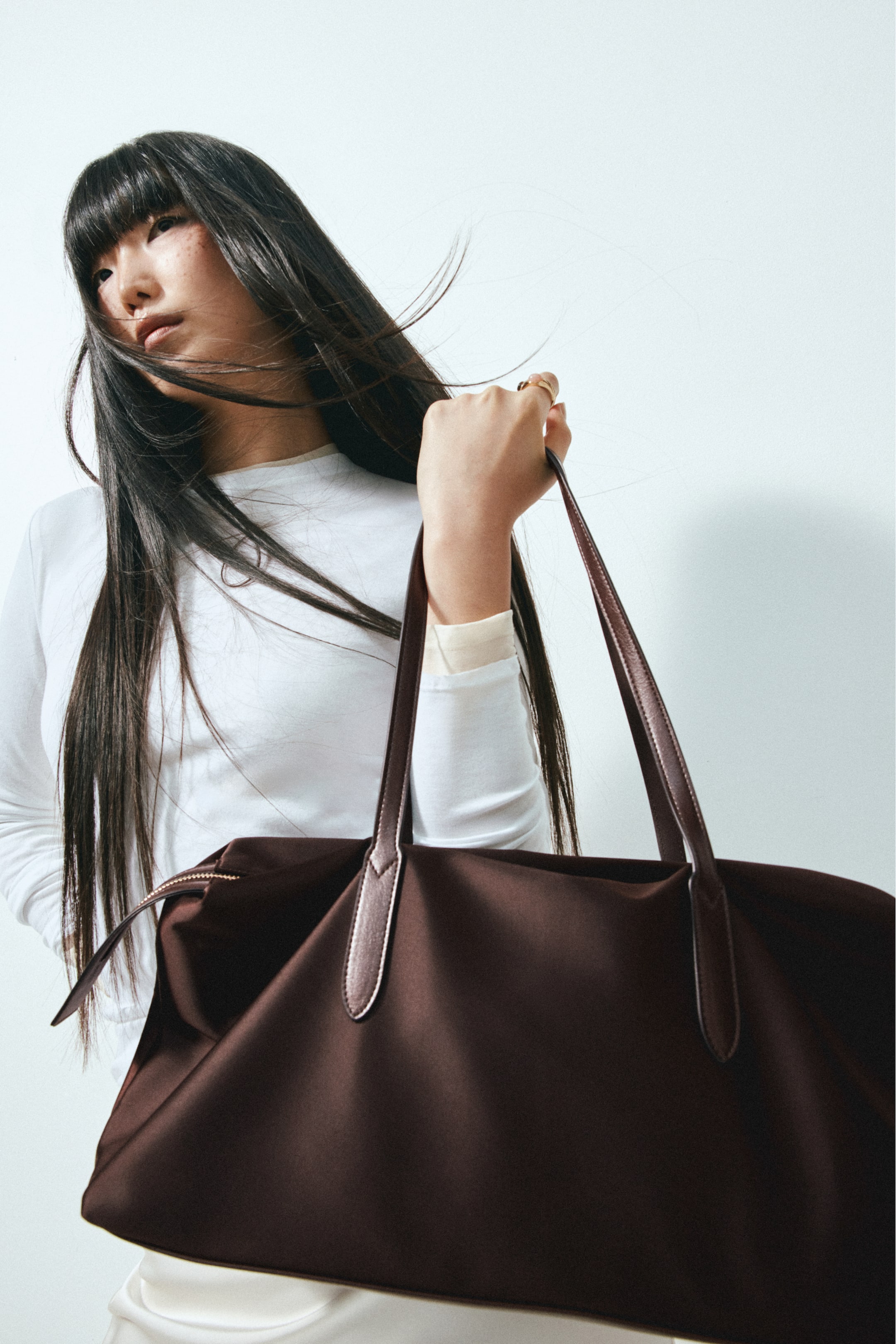 View larger image: A large dark brown shopper bag in a smooth woven fabric is being carried by the model, who is holding its long, narrow handles. The bag features a top zip closure with a gold-toned zipper and is styled with a long-sleeved white top.