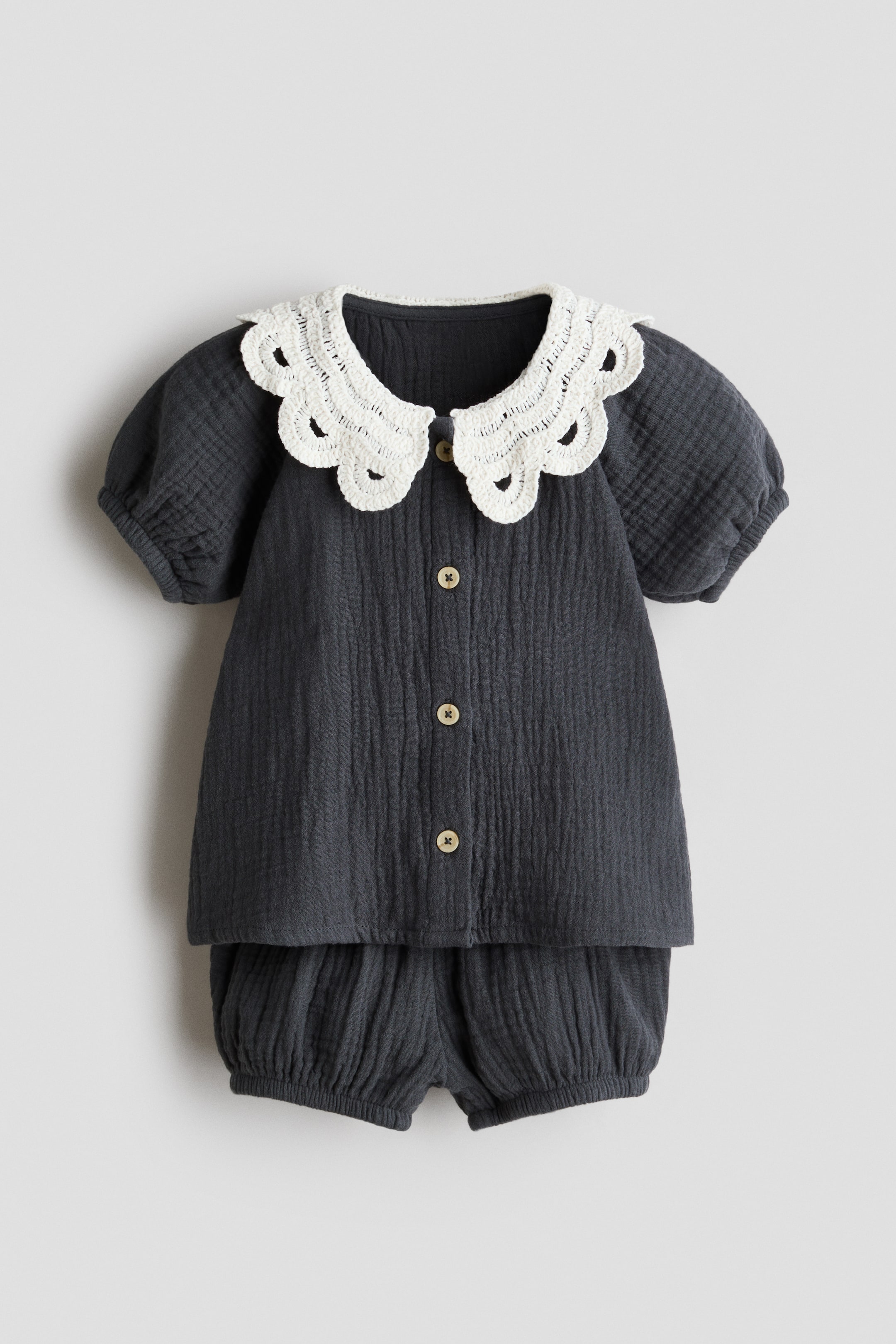 View larger image: A two-piece baby outfit featuring a dark grey textured top with a white, scalloped, crocheted Peter Pan collar, short puff sleeves, and a front button placket with three light brown buttons. The matching dark grey bloomers have elasticated leg openings, creating a gathered effect.