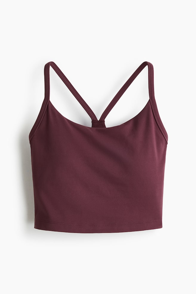 A maroon sports bra is presented from the front, featuring a scoop neckline and thin spaghetti straps that cross at the back. The smooth, matte fabric is forming a cropped length with a straight hem. - 1