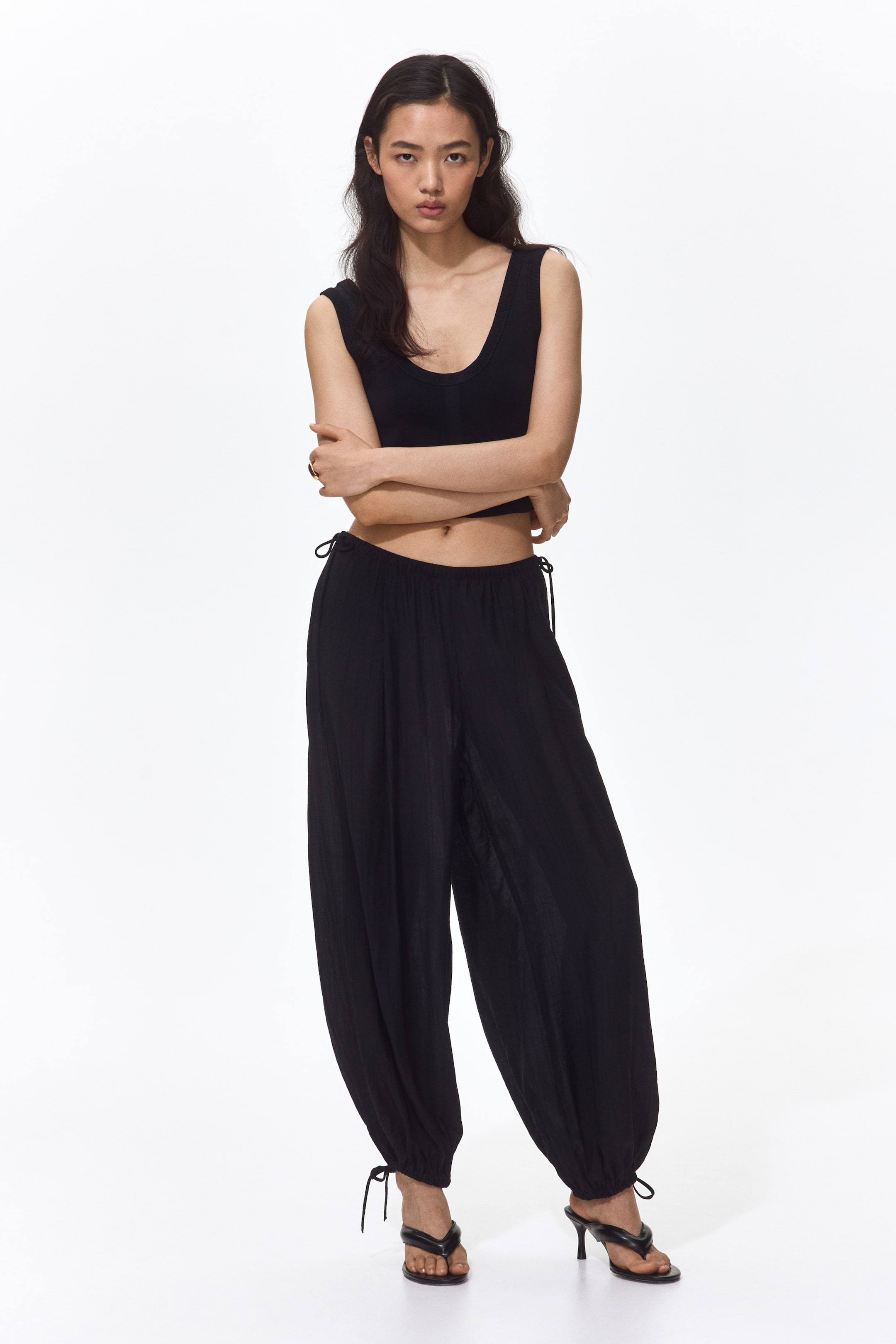 View larger image: A black sleeveless crop top featuring a deep scoop neck and a fitted silhouette. Black crinkled trousers showcase a high drawstring waist and a loose, billowing fit, gathering at the ankles with tied drawstrings.