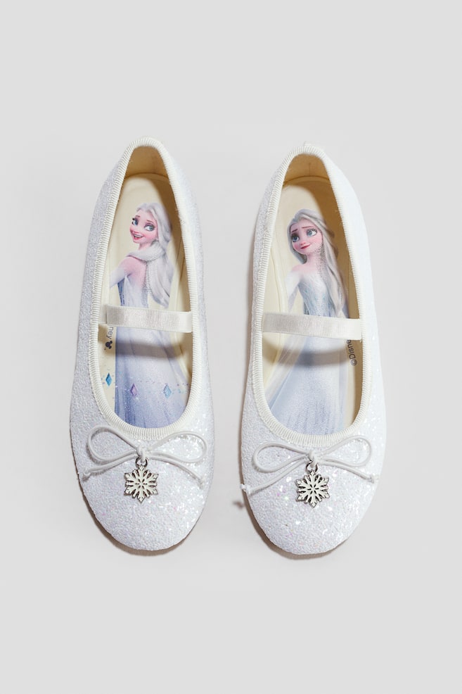 Ein Paar weiÃe, glitzernde Ballerinas ist in der Draufsicht zu sehen. Sie verfügen über einen elastischen Riemen über dem Spann sowie eine dekorative Schleife mit einem baumelnden Schneeflocken-Anhänger auf der Vorderseite. Die Innensohlen zeigen eine gedruckte Grafikillustration von Elsa aus Disneys Frozen. - 1