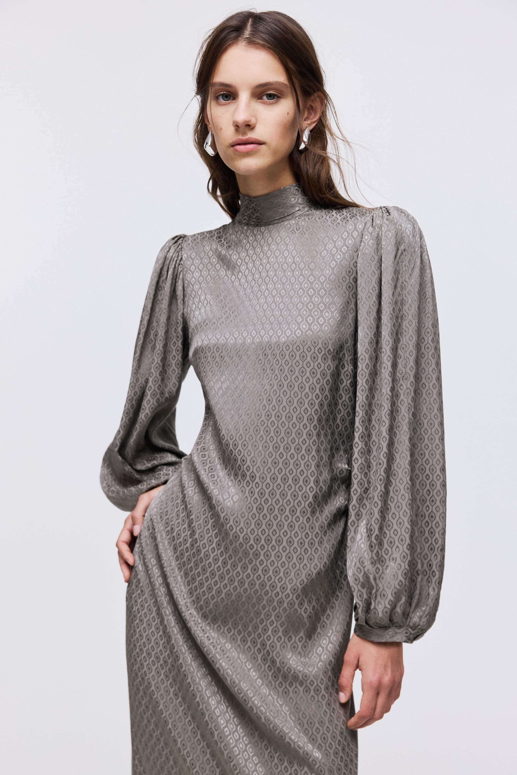 Se større billede: En knælang, tætsiddende kjole i grå viskosesatin med et subtilt geometrisk mønster bæres af modellen. Den har en standkrave og lange ballonærmer med smalle manchetter; modellen placerer den ene hånd på sin hofte.