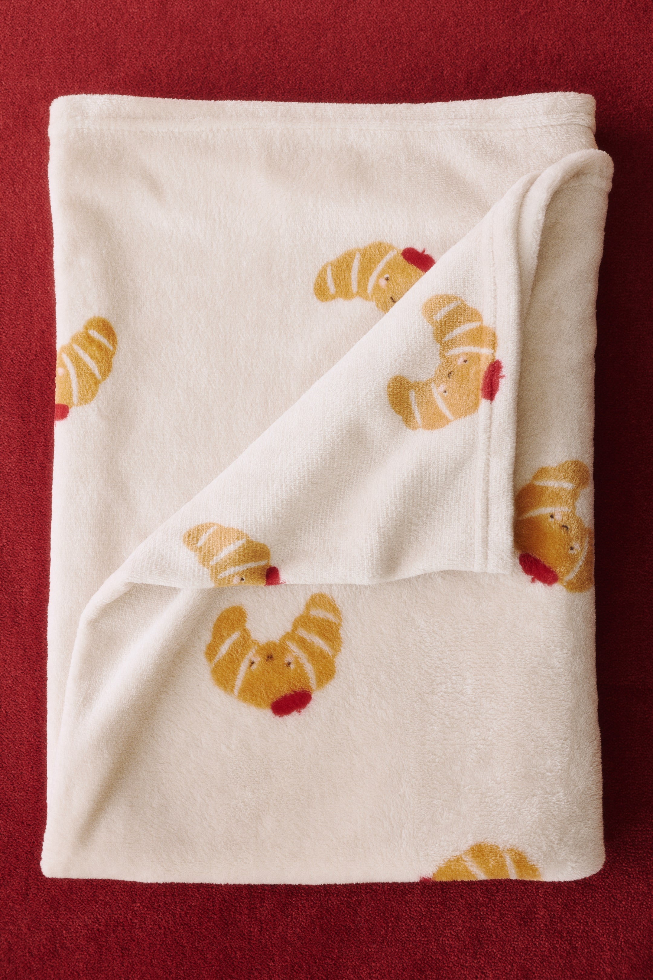 View larger image: A creamy white fleece blanket is lying folded, with one corner turned back, showcasing a repeated pattern of golden-brown croissants with small red accents.