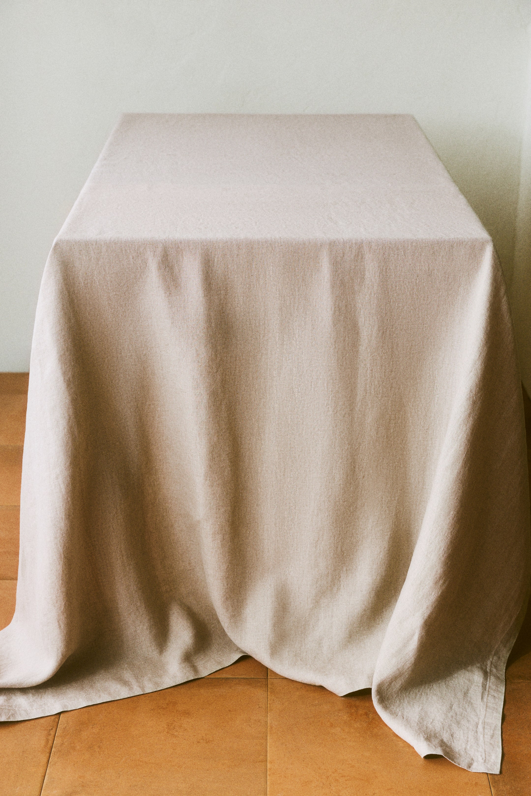 Grotere afbeelding bekijken: Een lichtbeige tafellaken hangt sierlijk over een rechthoekige tafel. Het toont een zichtbare linnen weefstructuur en een netjes gevouwen zoom langs de onderrand.