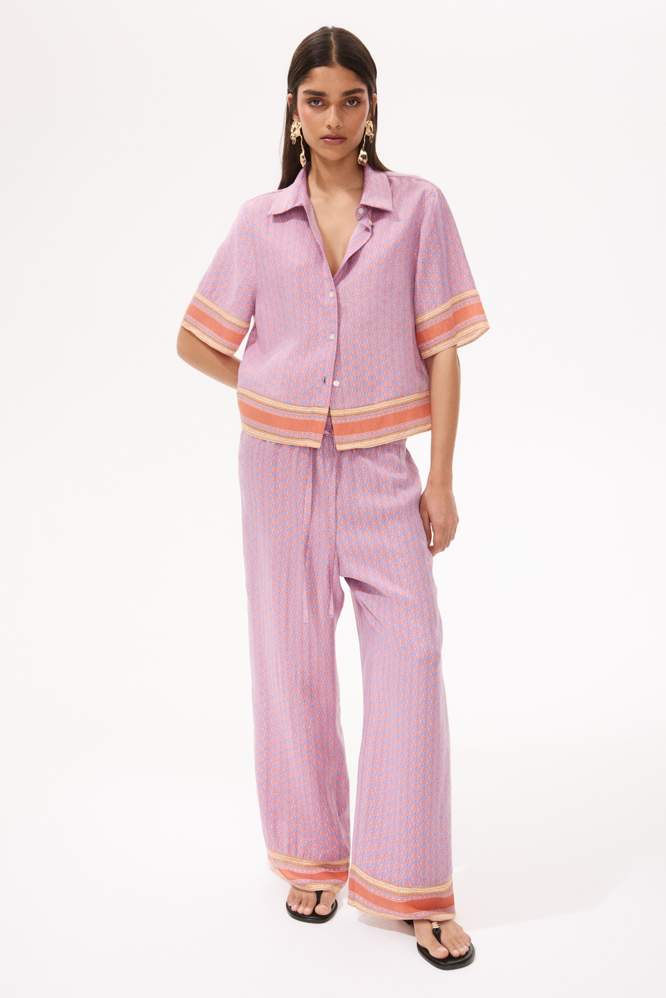 View larger image: This two-piece set includes a short-sleeved, collared button-up shirt and wide-leg trousers, styled to face forward. Both garments showcase a light purple and pink geometric design, highlighted with orange and yellow striped borders on the sleeve cuffs, shirt hem, and trouser hems. The ensemble offers a relaxed fit, with the shirt designed to be worn untucked.