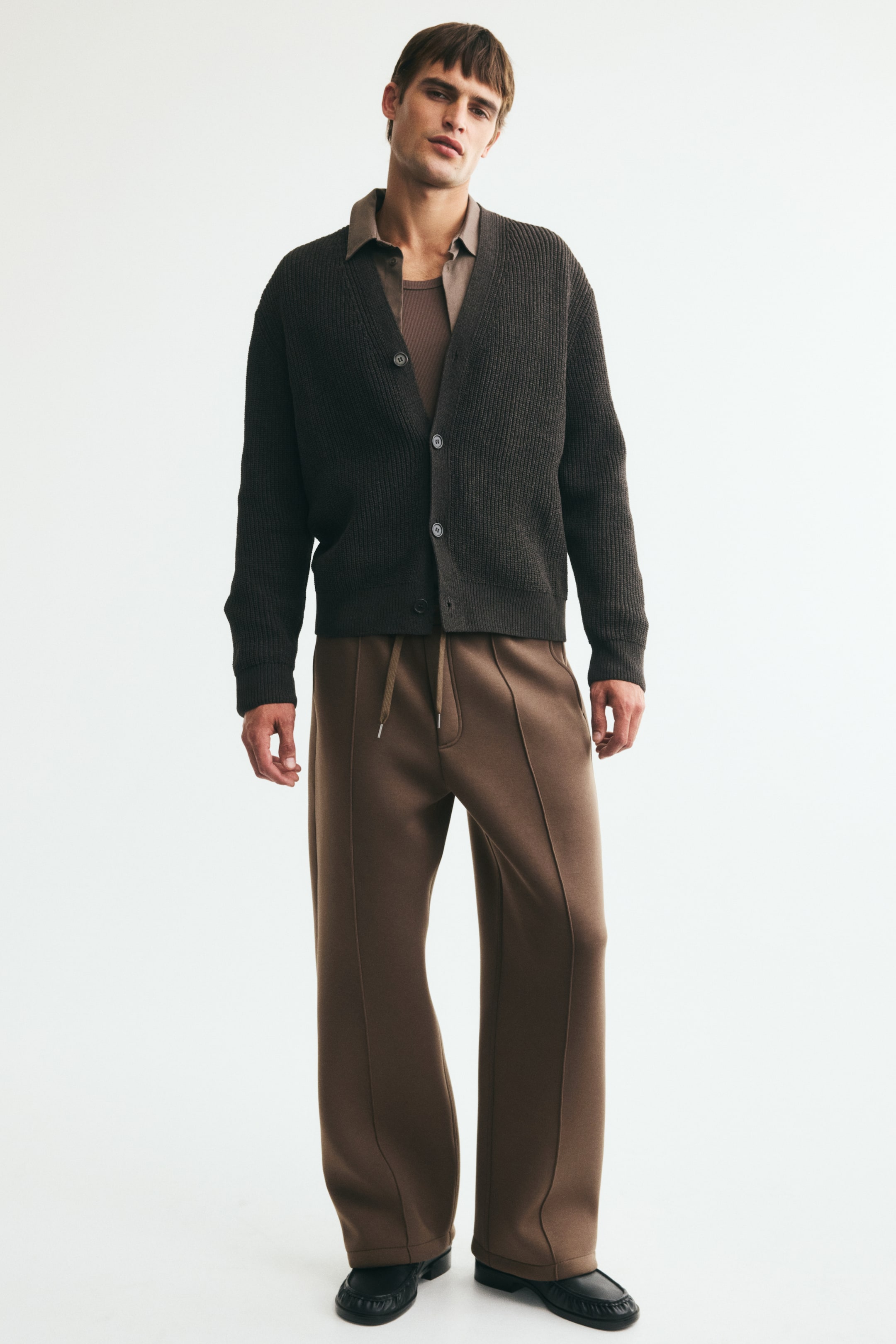 View larger image: Brown joggers in scuba fabric, featuring sewn-in creases down the front, an elasticized drawstring waist, and a faux fly, are worn by the model, showing a relaxed fit.