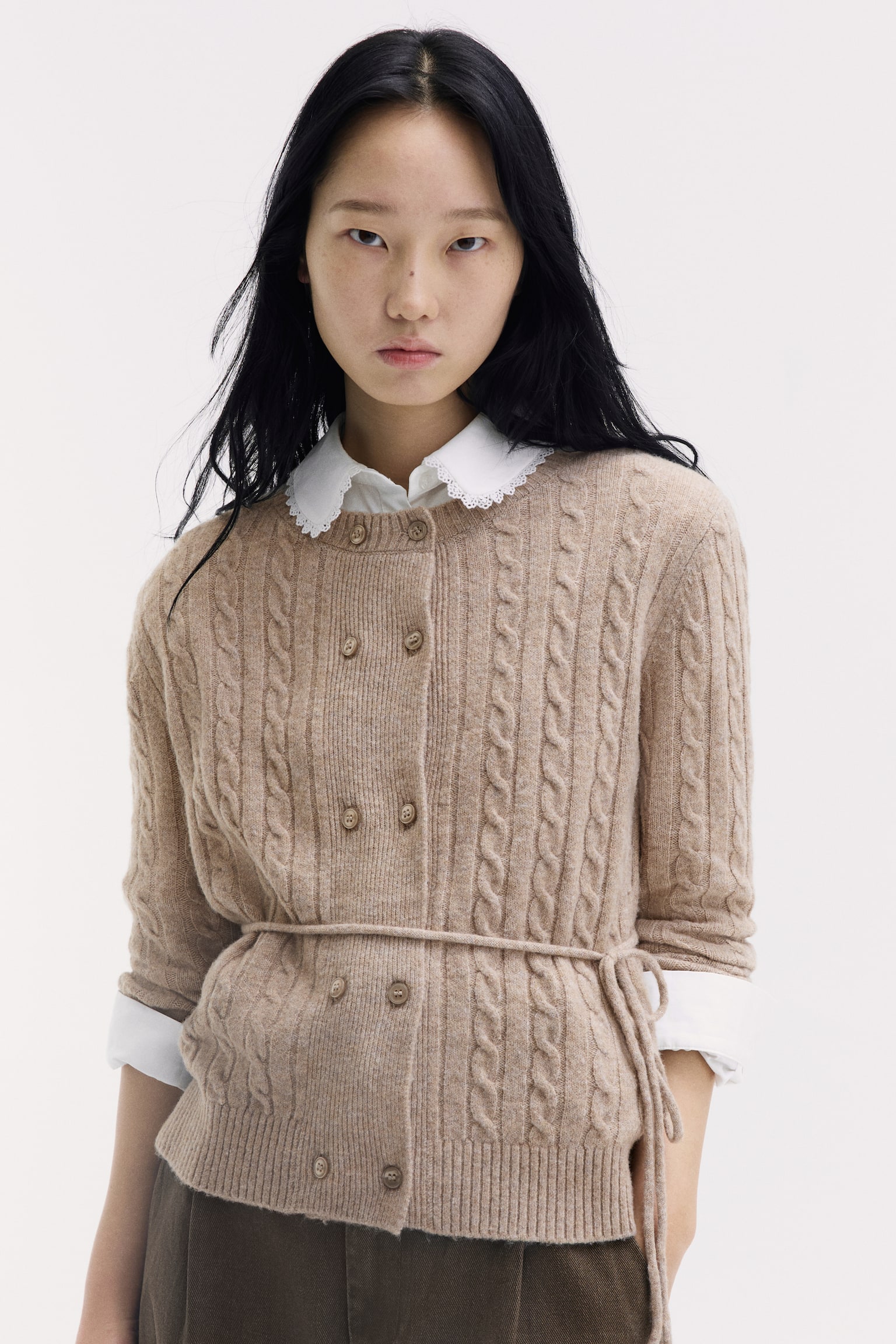 A beige cable-knit cardigan with a round ribbed neckline, double-breasted button closures, and a narrow waist tie belt is worn by the model. The cardigan is layered over a white shirt with a scalloped collar and paired with dark trousers. - 1