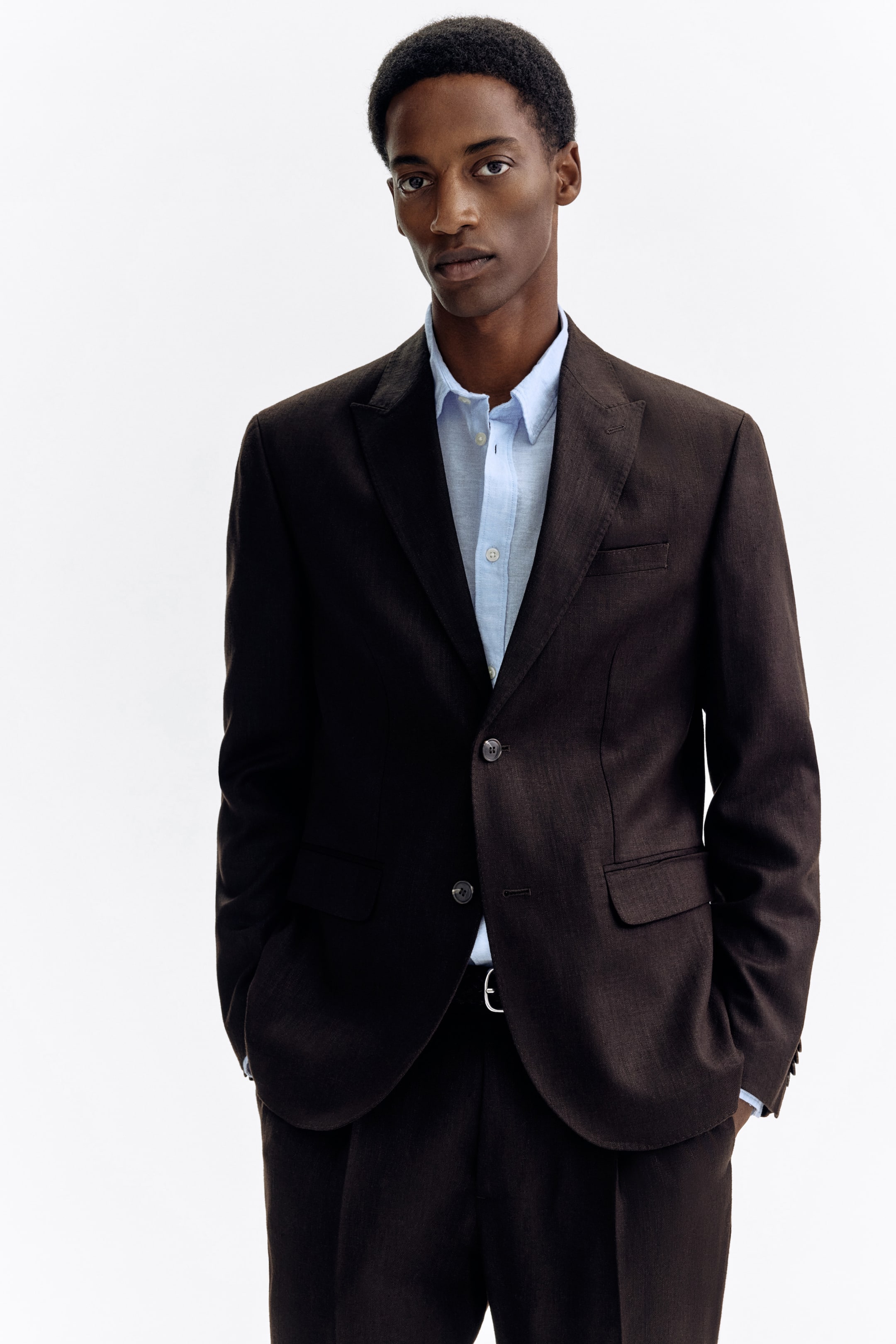 View larger image: A dark brown suit is being presented, facing slightly to the viewer's right, featuring a single-breasted jacket with notch lapels, a two-button closure, a chest welt pocket, and front flap pockets, layered over a light blue collared shirt. Matching dark brown trousers with pleated fronts complete the look, with a belt visible at the waist.