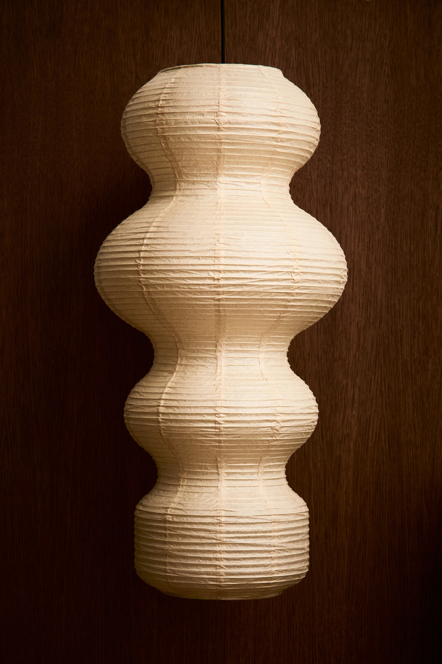 A pendant lamp featuring a textured cream-colored rice paper shade is hanging vertically, shown from a front angle. The shade showcases a sculptural, multi-tiered bulbous silhouette with horizontal ribbing and is softly glowing from an internal light source. - 1