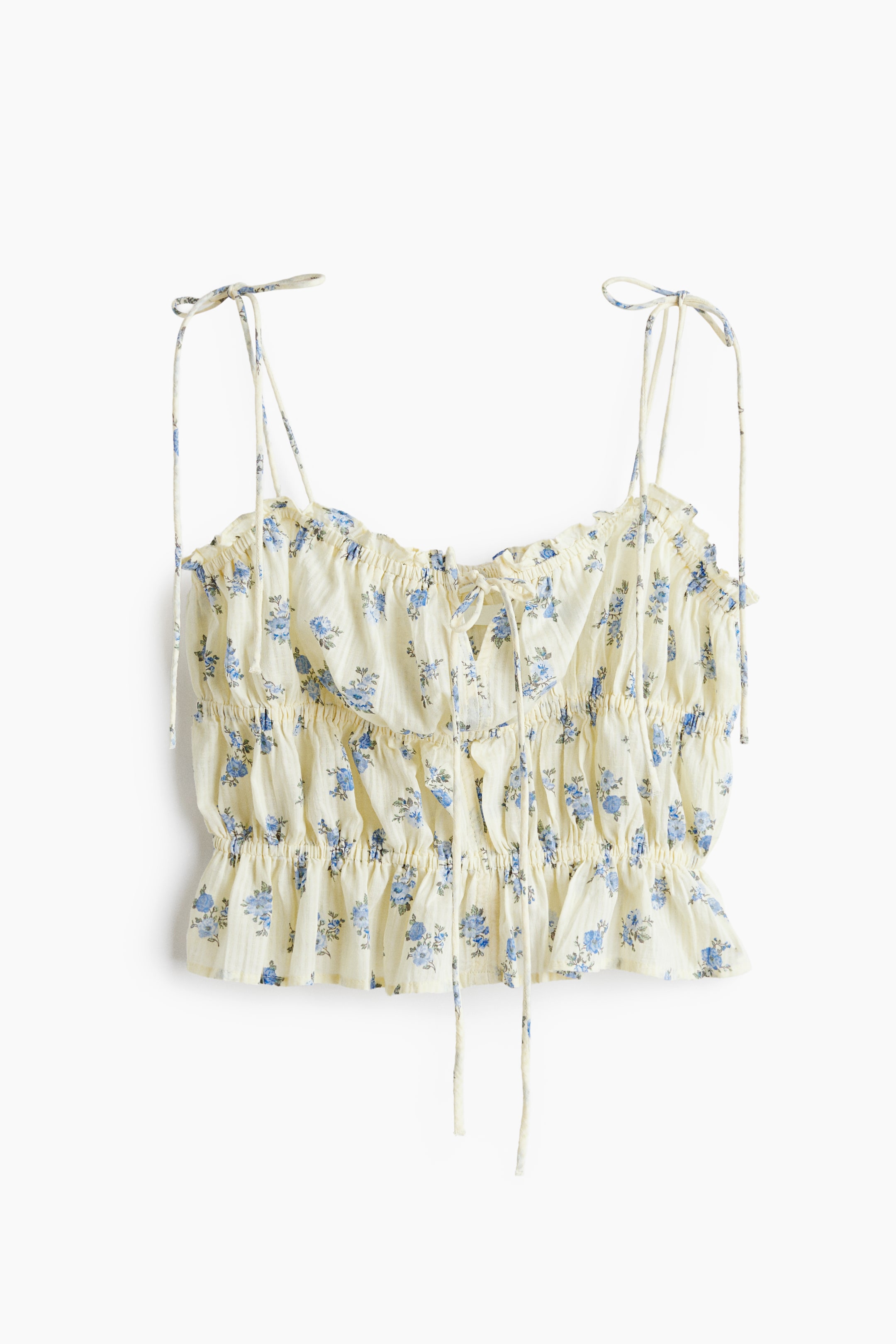 View larger image: The cream crop top showcases a blue floral pattern and highlights smocked detailing across the front, offering a gathered texture. It comes with tie-up spaghetti straps and a center front tie, complemented by a ruffled hemline.