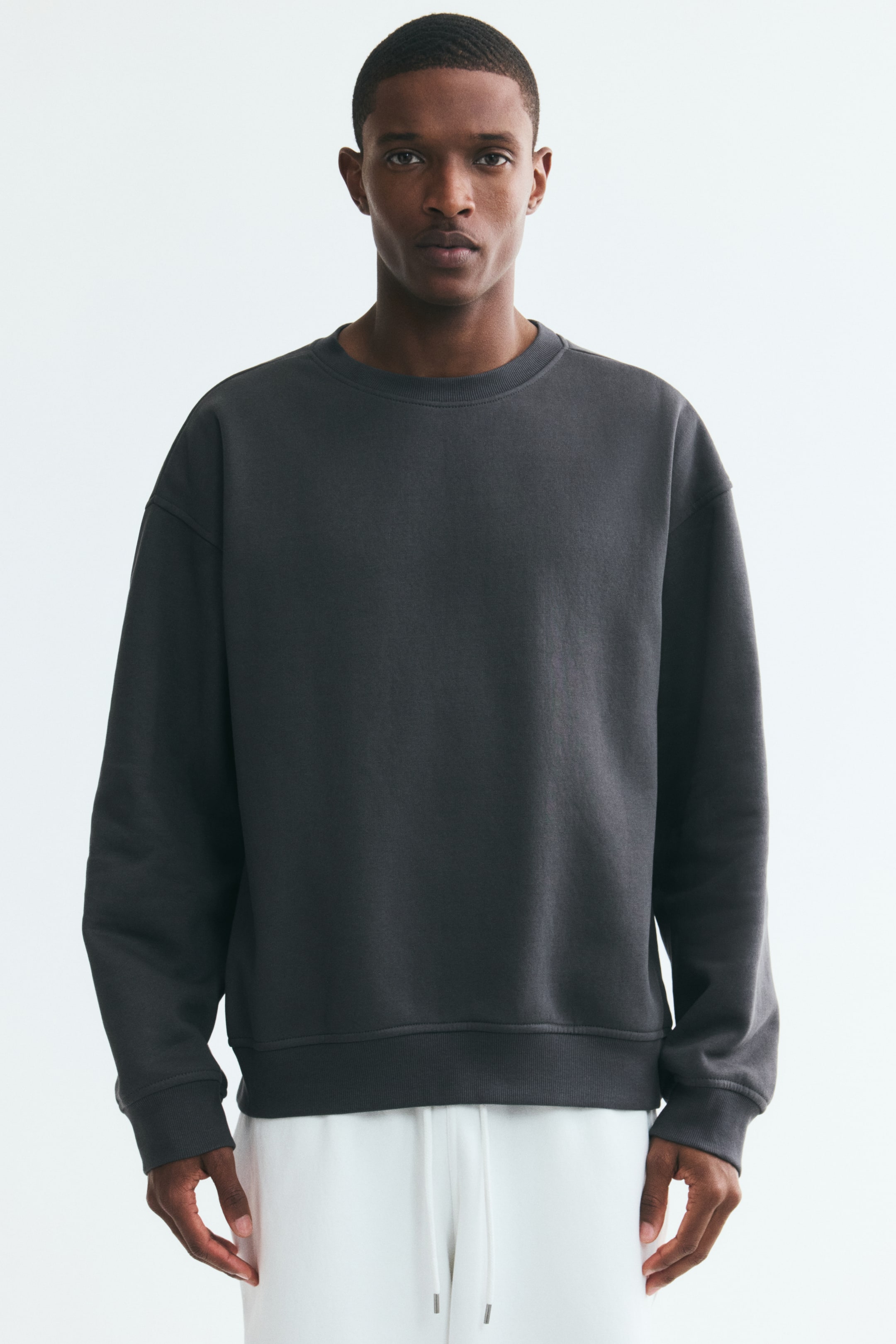 View larger image: A dark grey sweatshirt featuring a round, rib-trimmed neckline, dropped shoulders, long sleeves with wide ribbed cuffs, and a wide ribbed hem is being worn by the model. The model is styling the loose-fit sweatshirt with white pants.