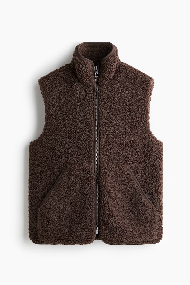 A brown sherpa fleece vest is being shown from the front. It features a full-length zipper closure, a stand-up collar, and two angled front pockets, all bordered with darker brown fabric binding. - 1