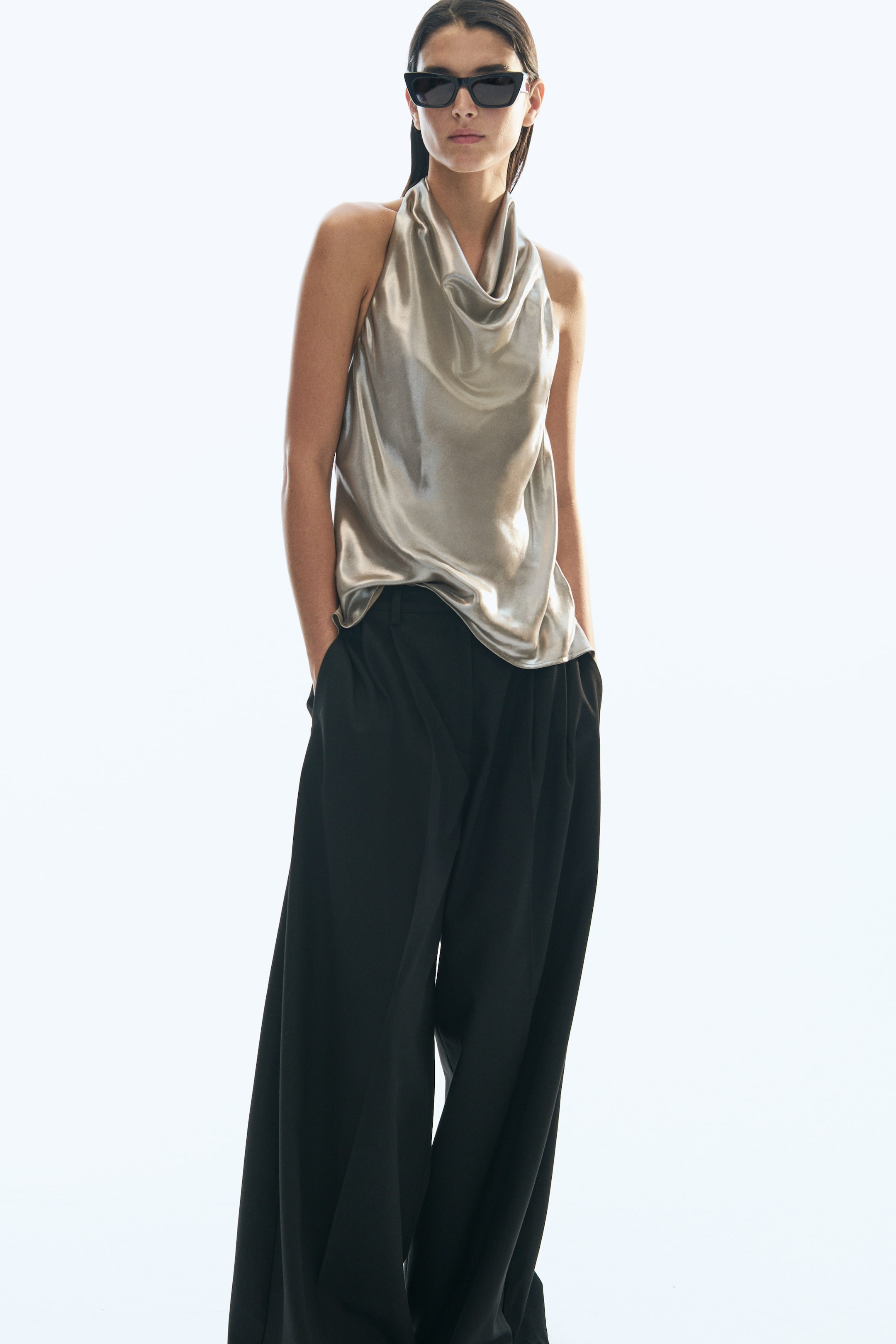 View larger image: A sleeveless, relaxed-fit top in draped champagne-coloured satin is worn by the model, featuring a cowl neckline and halterneck silhouette. The top is styled with wide-leg black trousers.