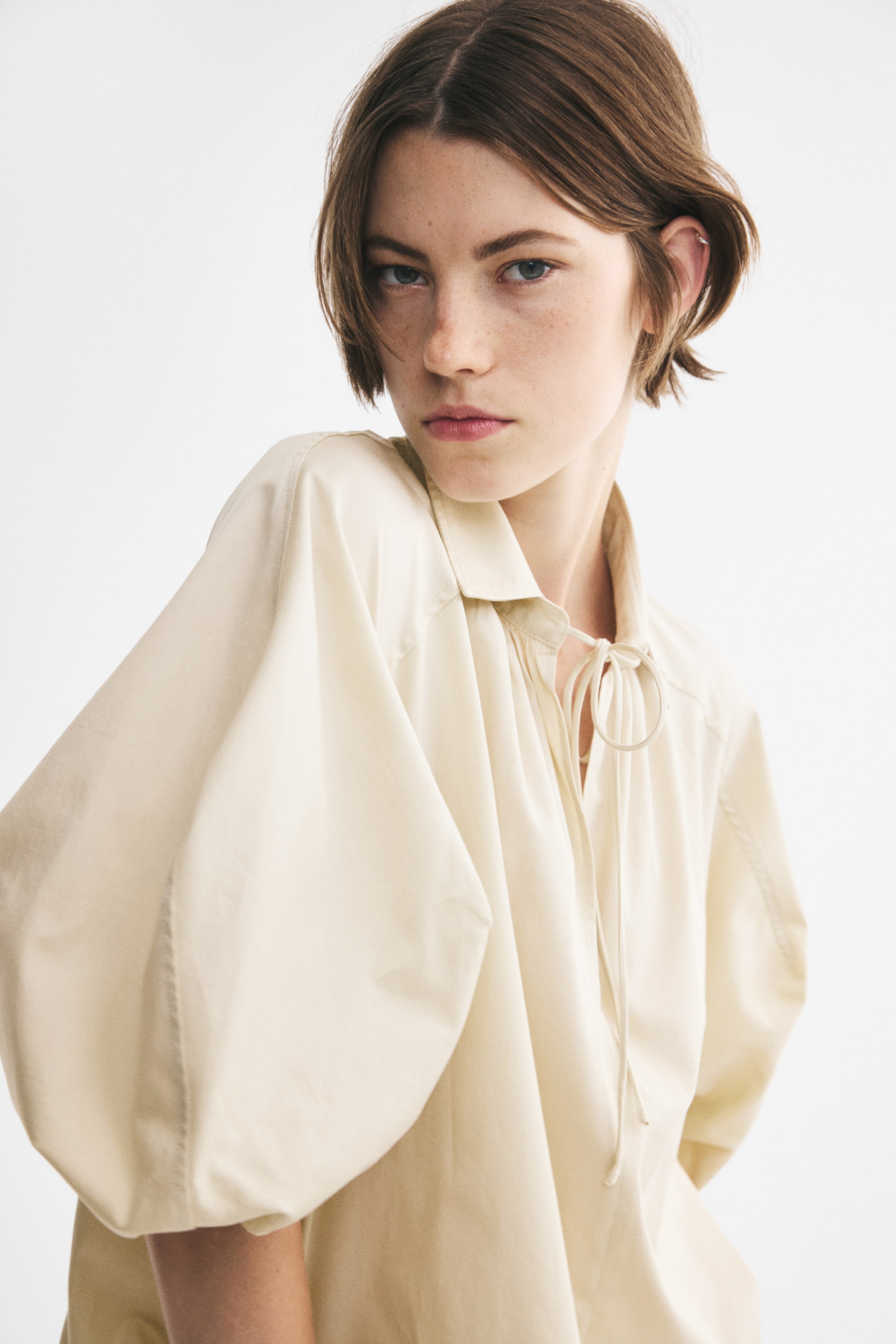 Agrandir l'image: Une blouse en popeline de coton couleur crème présente un col et une ouverture en V avec de longs liens étroits sur le devant. La blouse est dotée d’empiècements d’épaules façon cavalier et de manches bouffantes volumineuses, longueur au coude, avec poignets élastiqués, mettant en valeur sa structure nette vue de trois quarts face.