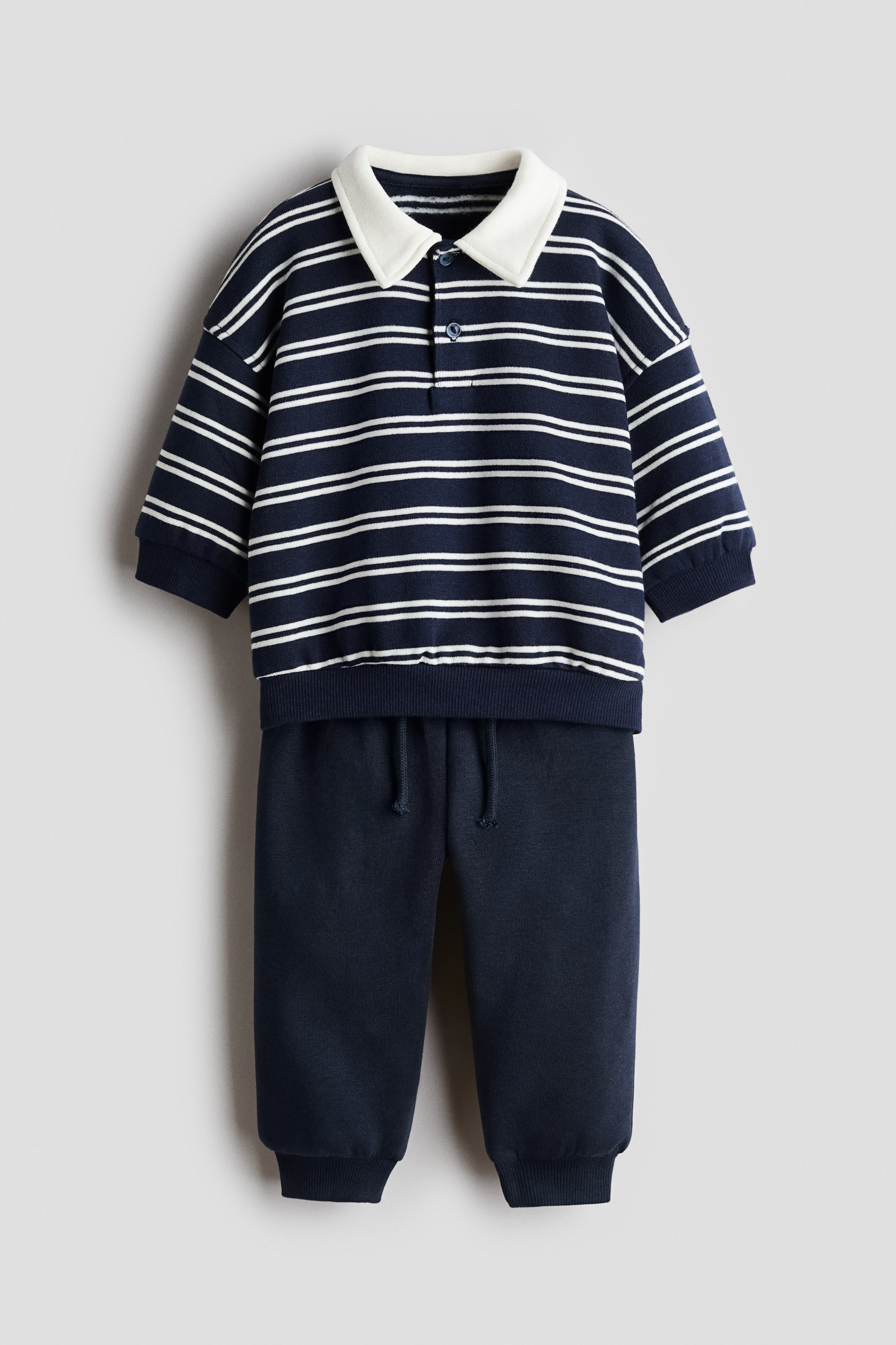 View larger image: A two-piece set, made of sweatshirt fabric, is being presented from the front, featuring a navy blue and white striped rugby shirt and solid navy blue joggers. The rugby shirt is designed with a white collar, button placket, dropped shoulders, long sleeves, and ribbed cuffs and hem, while the joggers are showcasing an elasticated waist with a decorative drawstring and ribbed hems.